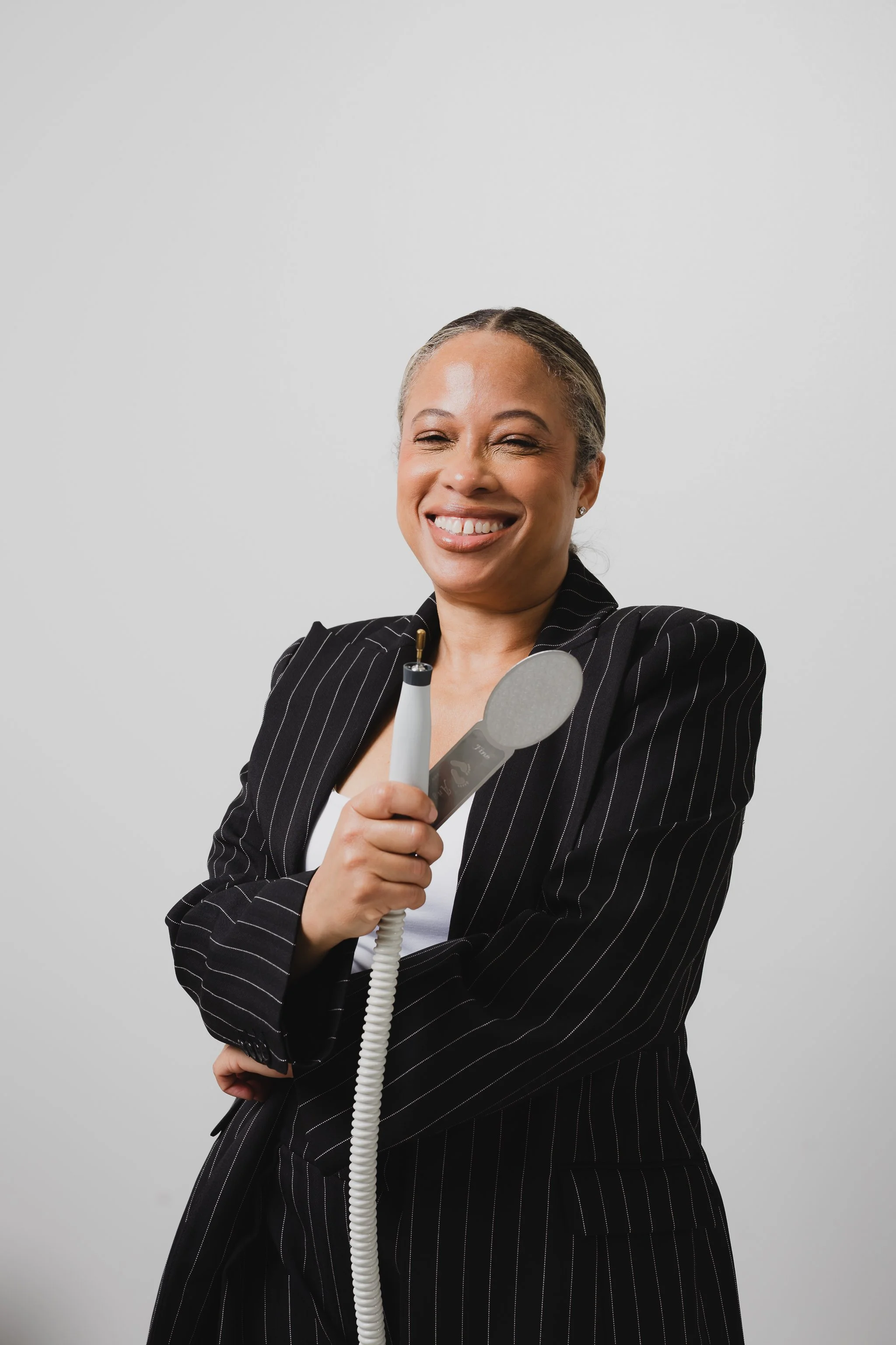A woman with blonde hair in a bun, smiling with closed eyes, wearing a black pinstripe suit and holding a telephone receiver connected to an old-fashioned corded phone, standing against a plain wall.