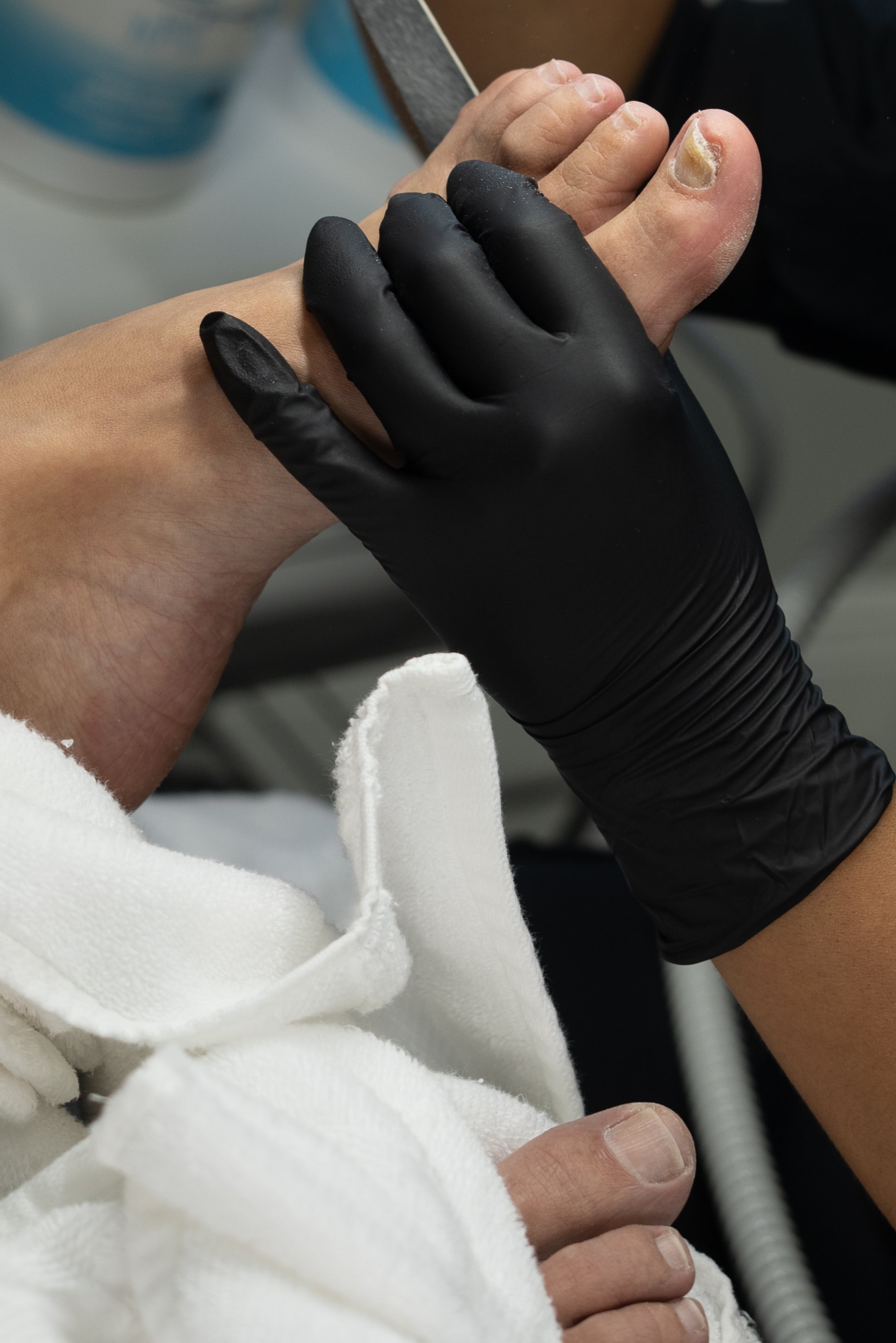 Close-up of a person's feet during a pedicure, with a technician wearing black gloves filing a toenail. The person's toenail is thickened and discolored. The person is wearing a white towel or robe.