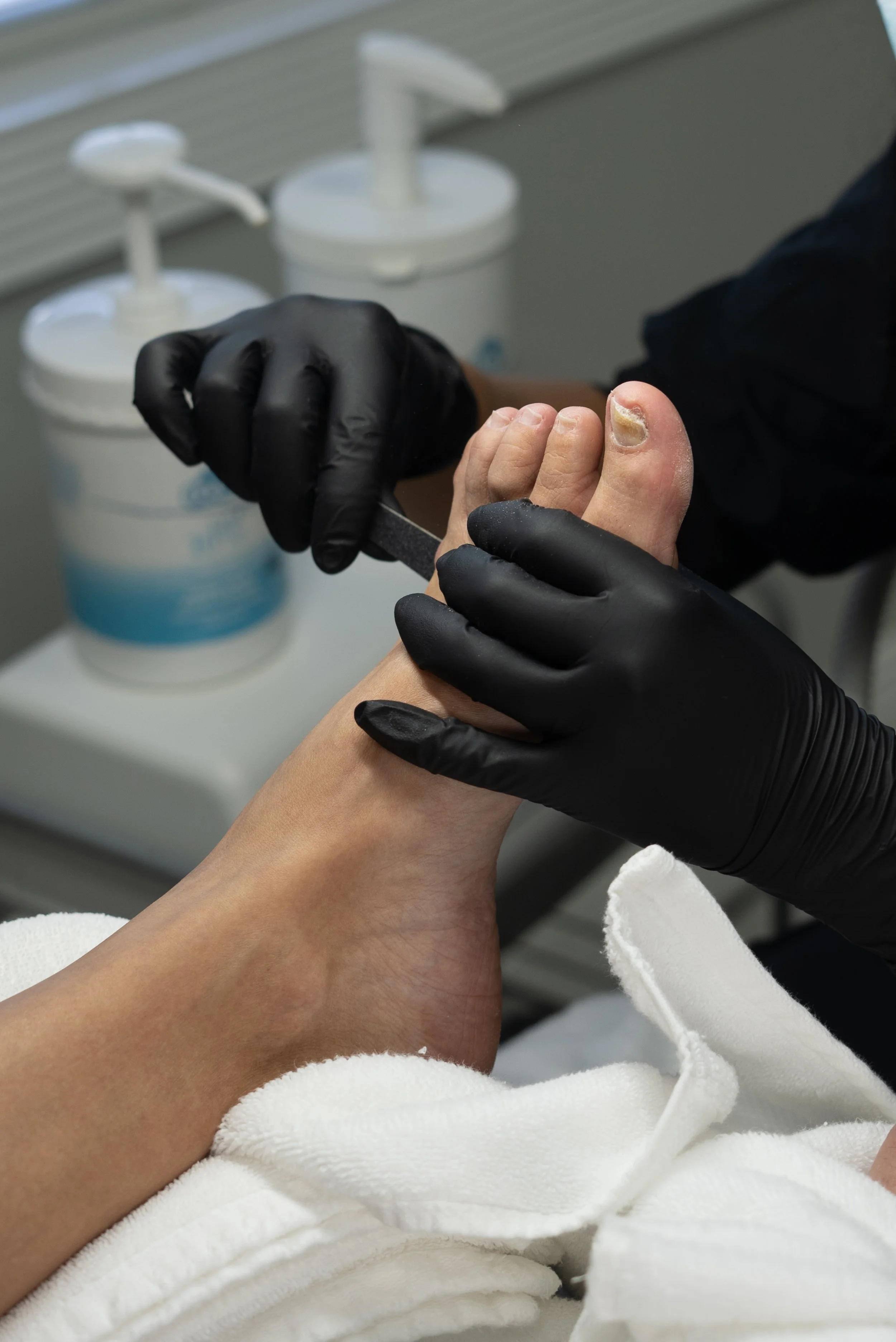 A person receiving a pedicure, with a foot being prepared for nail care by a technician wearing black gloves.