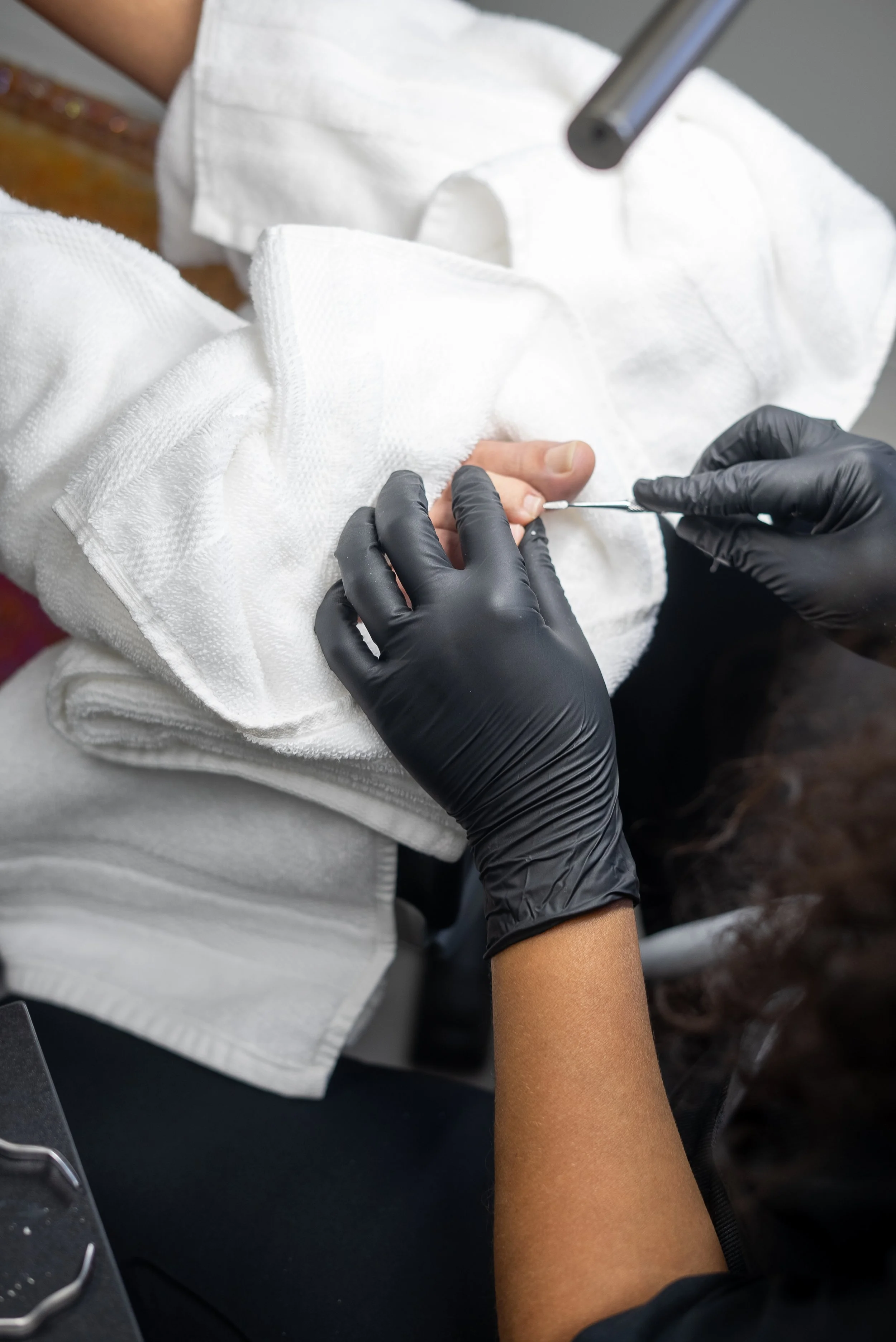 A person receiving a manicure, with another person wearing black gloves applying nail polish to their nails.