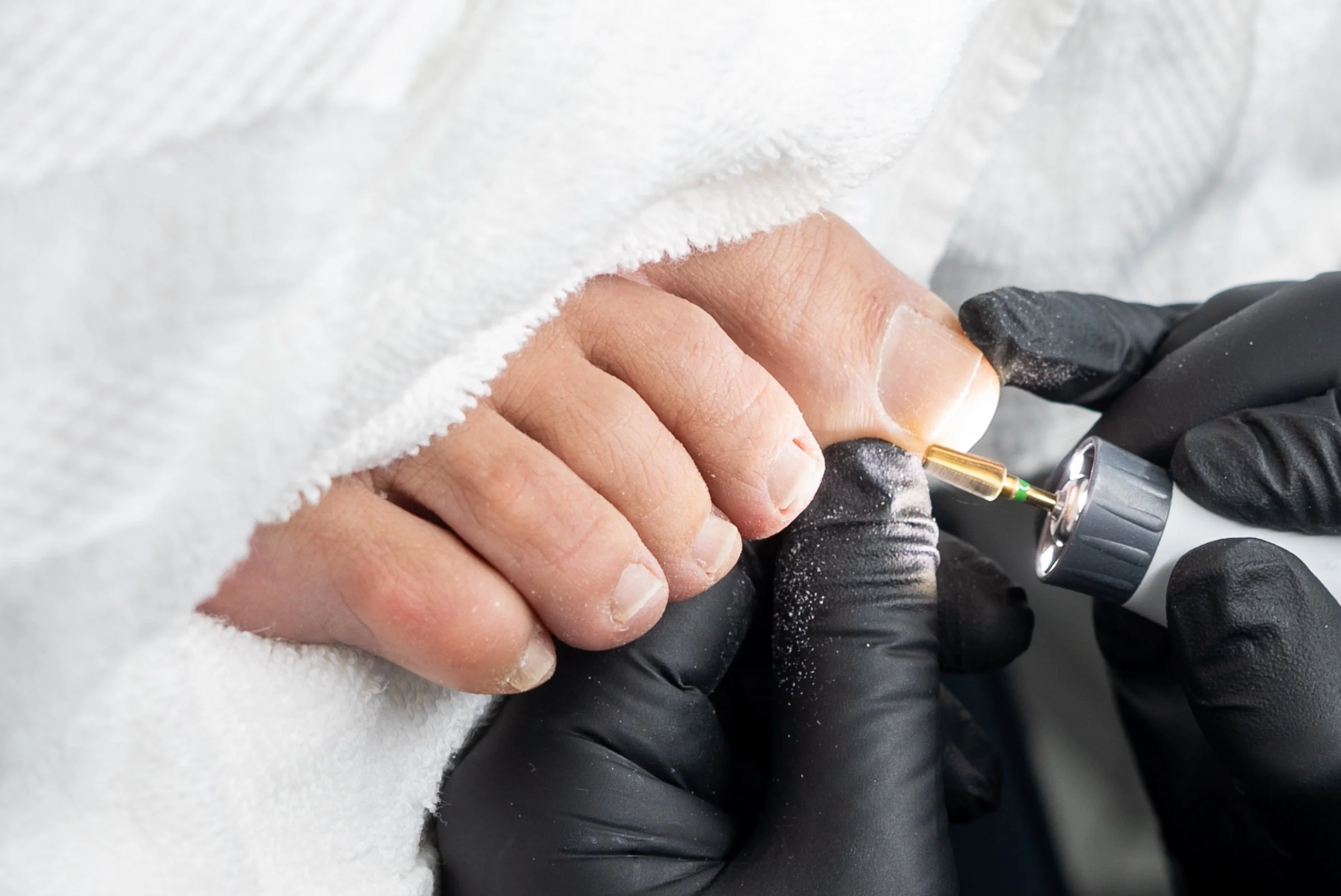 Close-up of a person receiving a manicure, with a nail technician using a tool for nail care while wearing black gloves, and the client's fingers resting on a white towel.