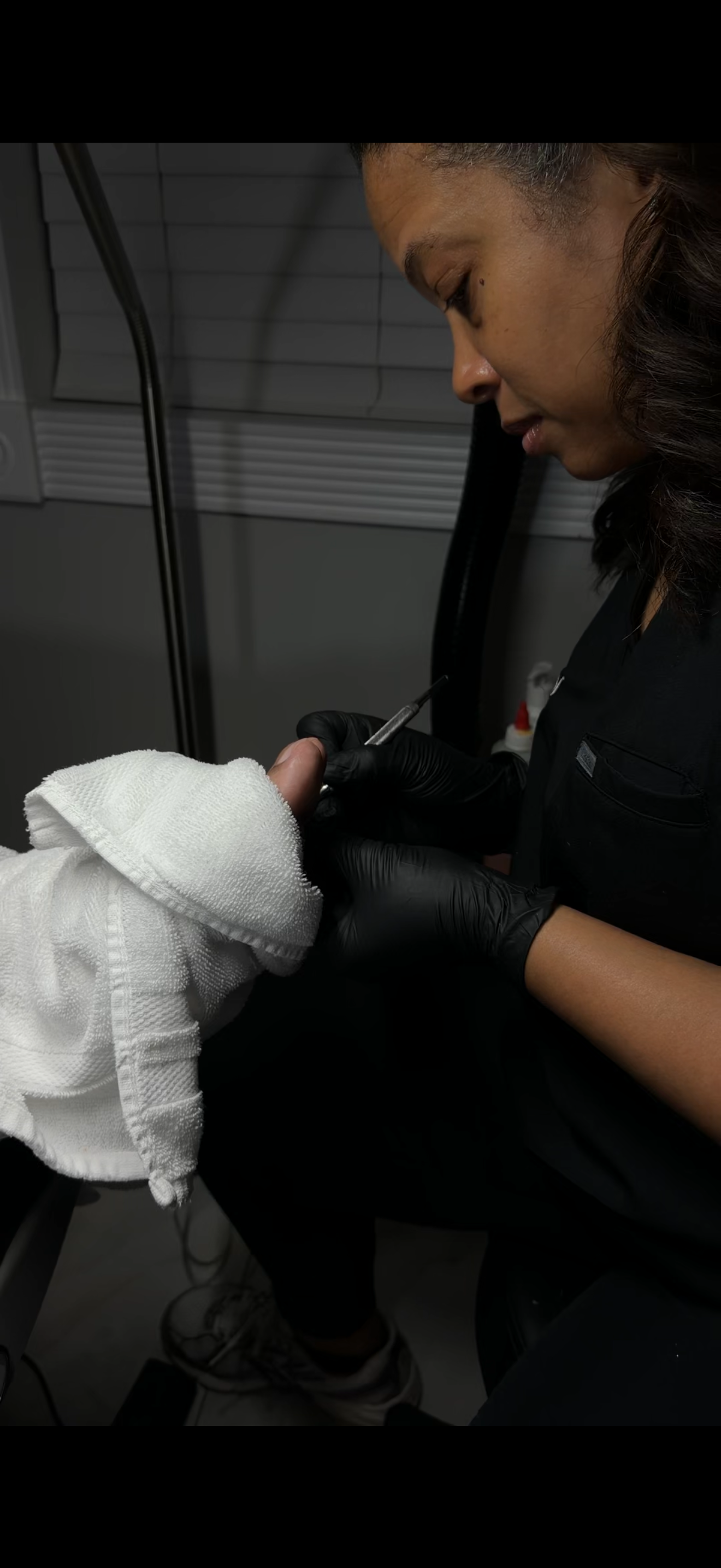 A woman with dark hair wearing black gloves administering a tattoo to a person's finger using a tattoo machine.