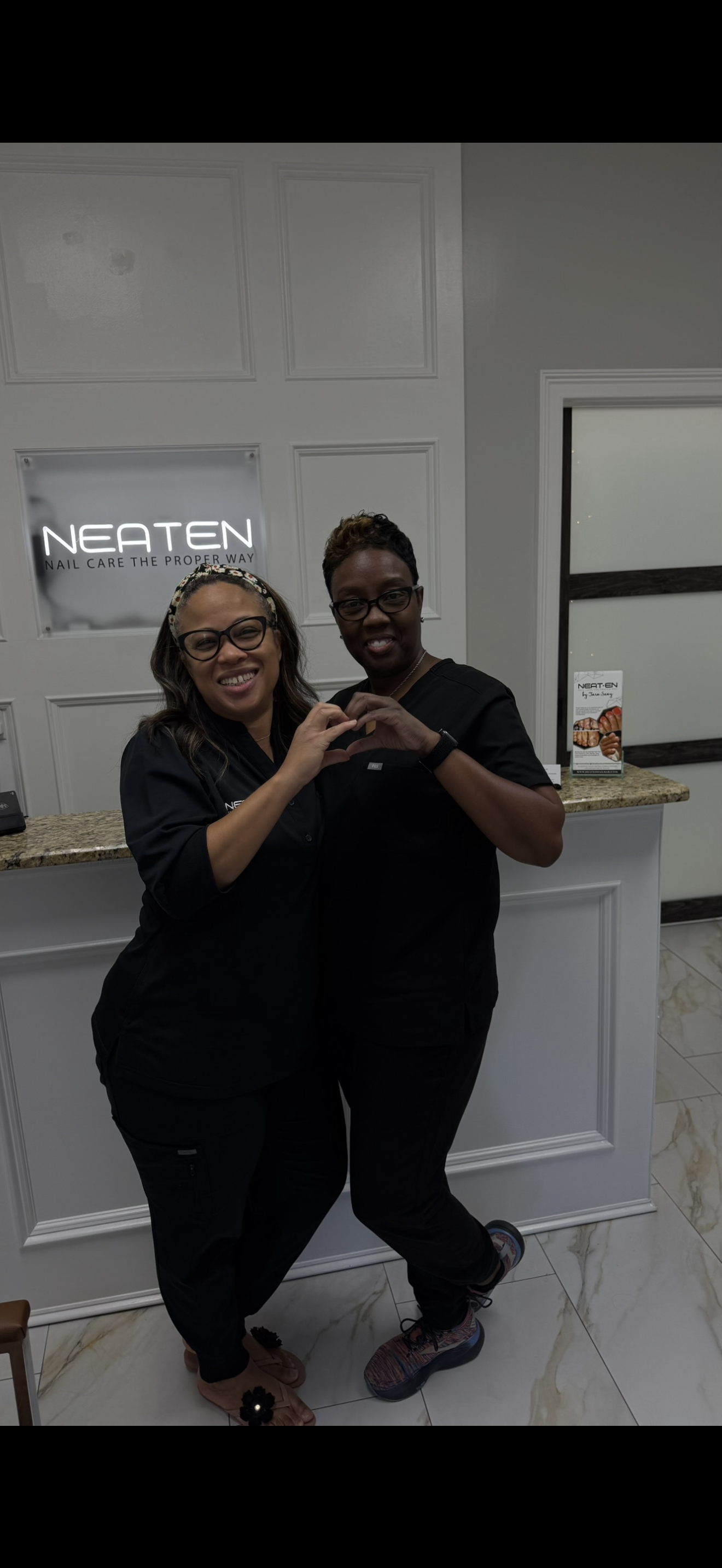 Two women standing in front of a reception desk, smiling and forming a heart shape with their hands. They are in a nail care salon, with a sign behind them that reads 'NEATEN Nail Care the Proper Way.'