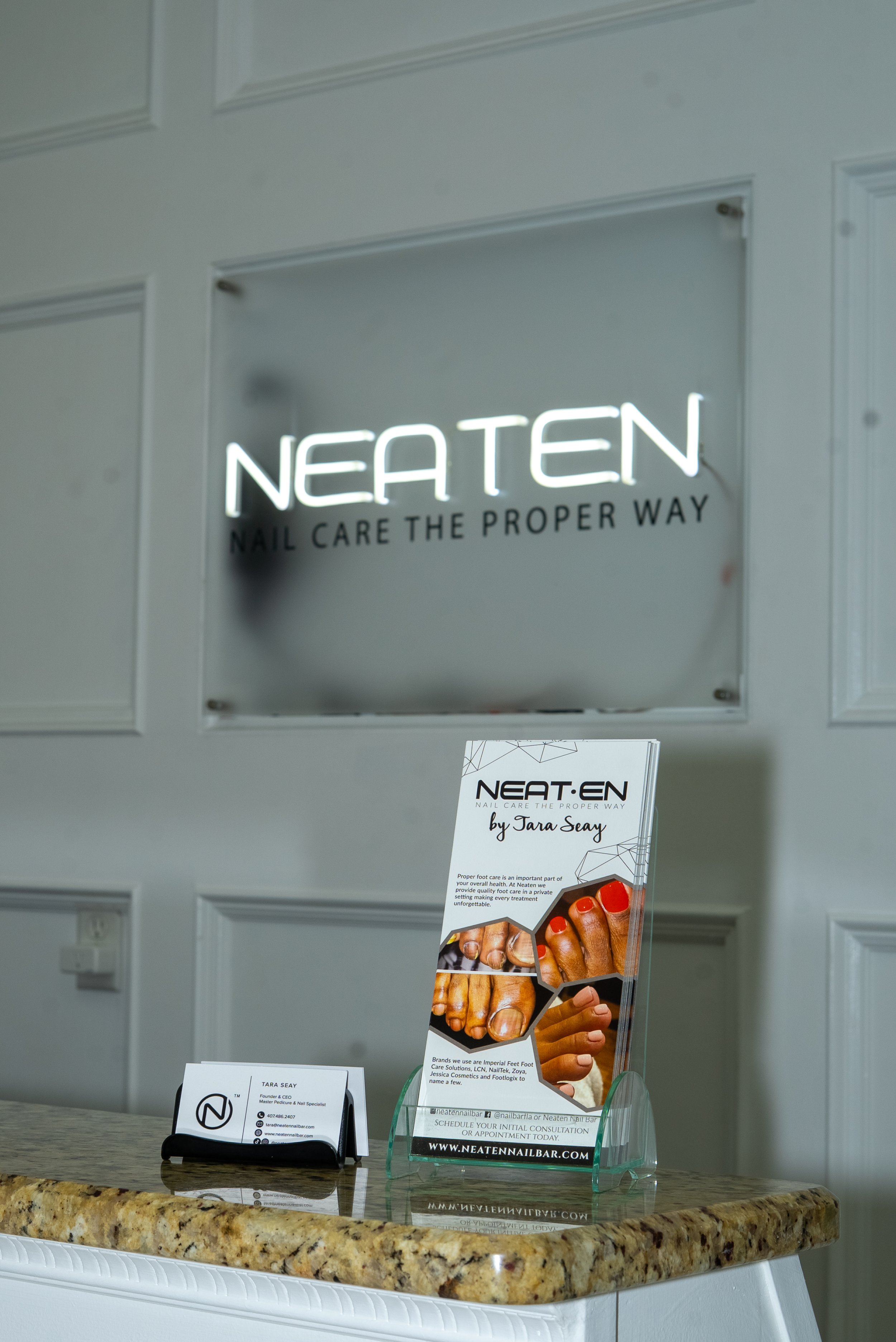 A neon sign that reads 'Neaten' with the slogan 'Nail Care The Proper Way' underneath it, mounted on a wall. Below the sign on a granite countertop, there is a brochure about nail care and a business card holder with a card that displays the name Tara Seay and contact information for Neaten Nail Bar.