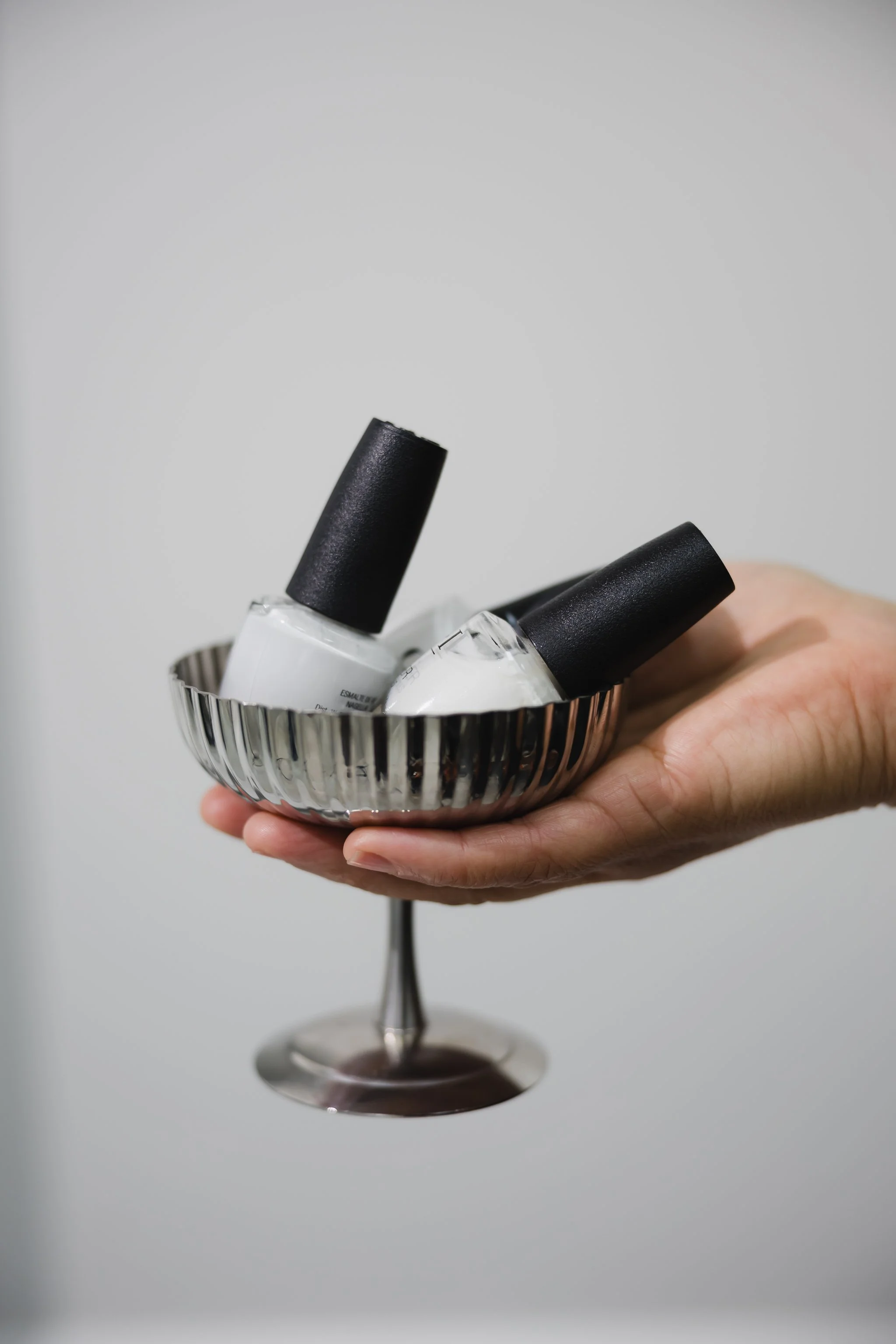A hand holding a silver goblet containing two bottles of white nail polish