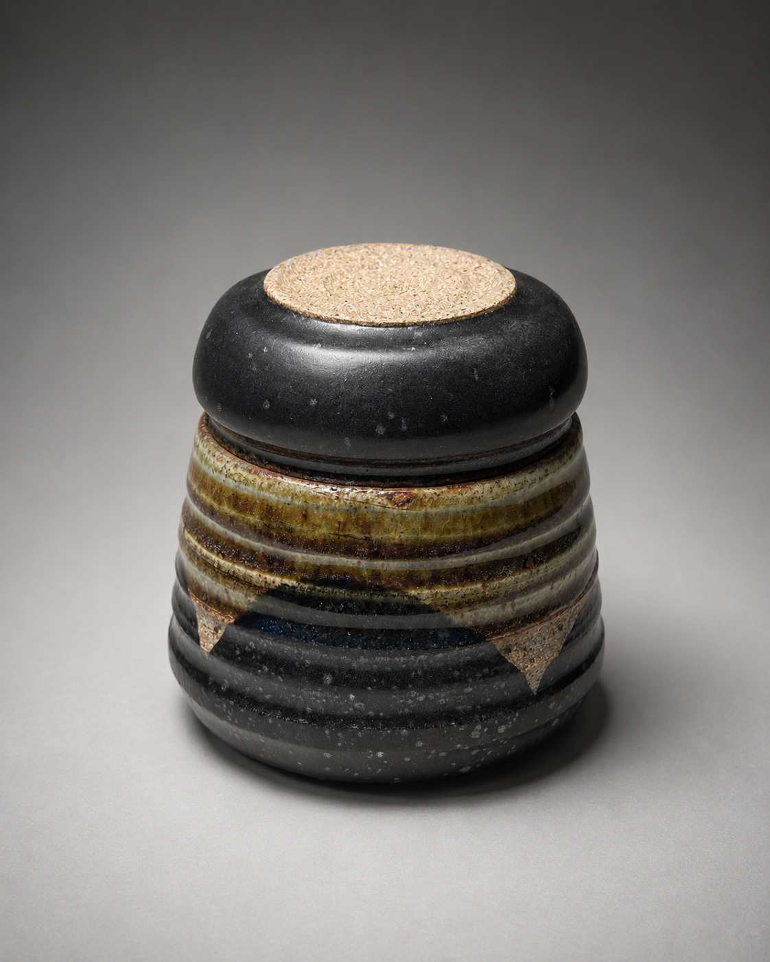 A ceramic or stone vessel with a rounded, ribbed design, featuring a black top, glazed, textured surface, and earthy tones, set against a neutral background.