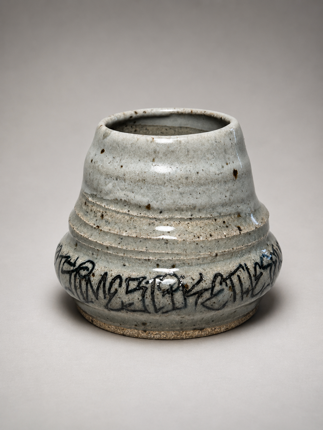A ceramic vase with a rounded shape, gray glaze, and black abstract markings around its midsection, set against a plain, neutral background.