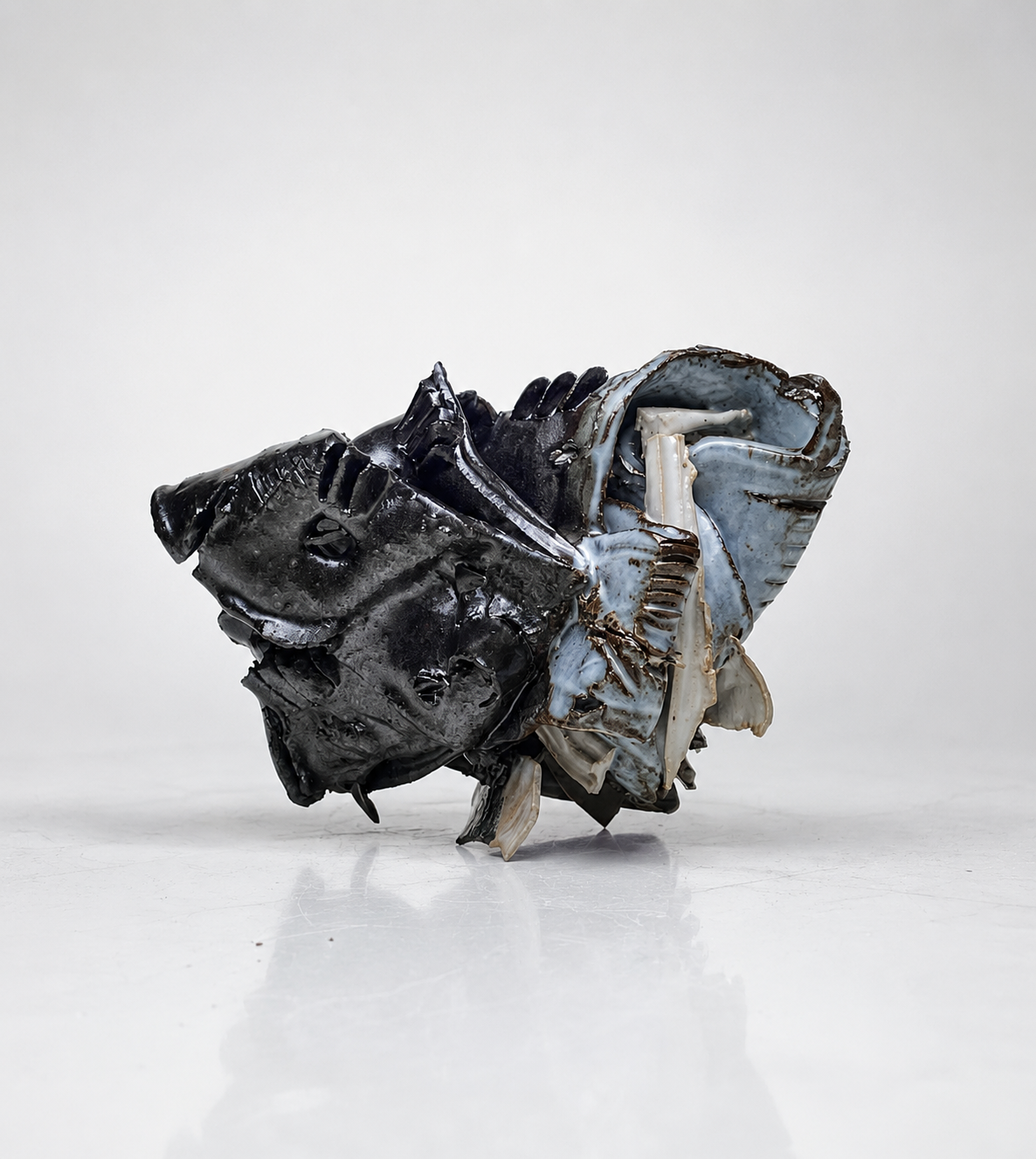 Sculpture made from crumpled, melted, and layered black and white plastic or metal materials, arranged into an abstract, organic shape on a white surface against a light background.