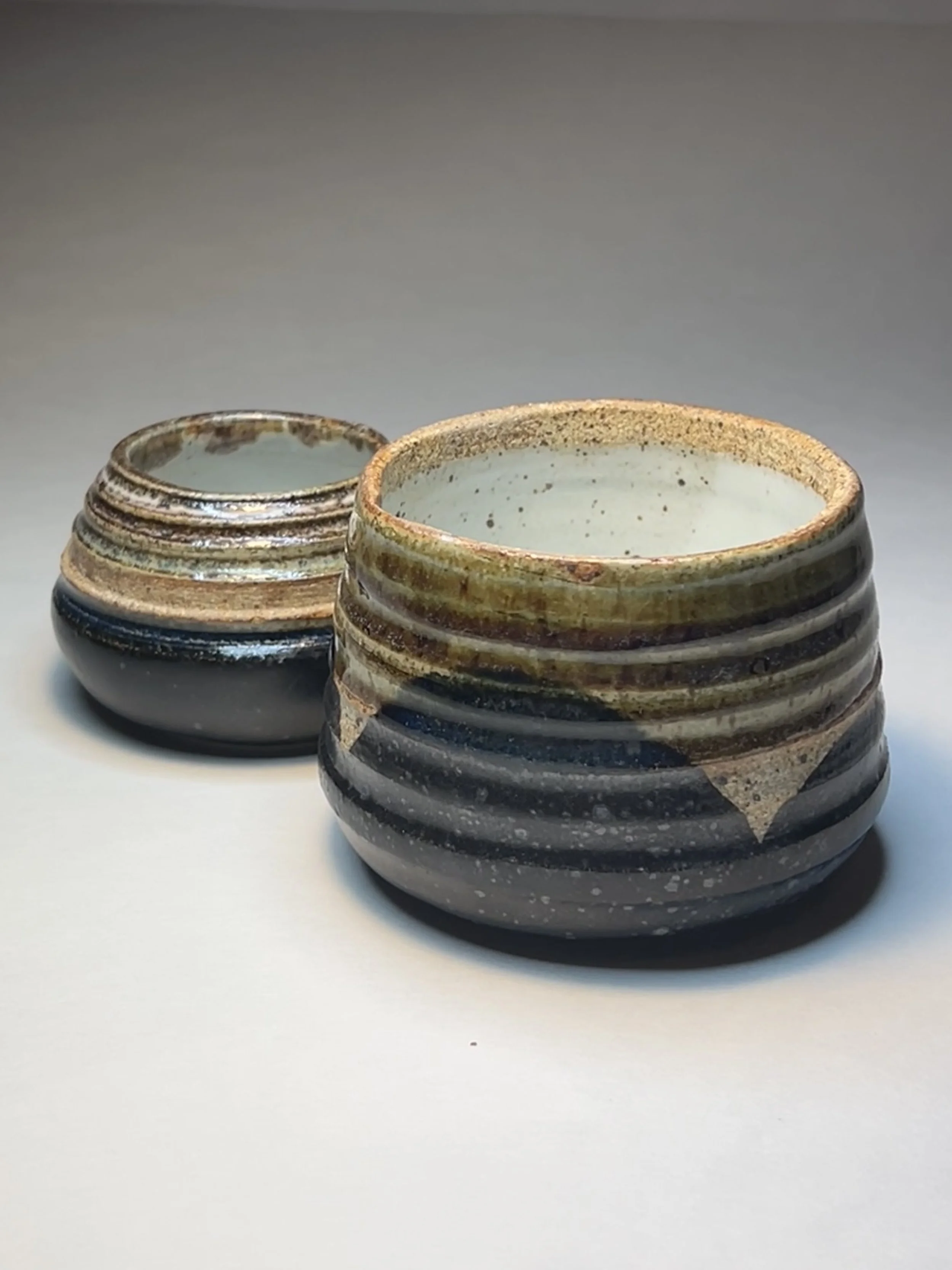 Two handcrafted ceramic pots with a rustic, earthy glaze, featuring horizontal ridges and speckled interior, placed on a plain white surface.