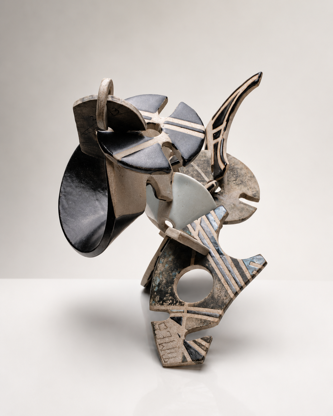 Abstract sculpture made of various geometric shapes and patterns, with earthy tones and black accents, set against a simple gray background.