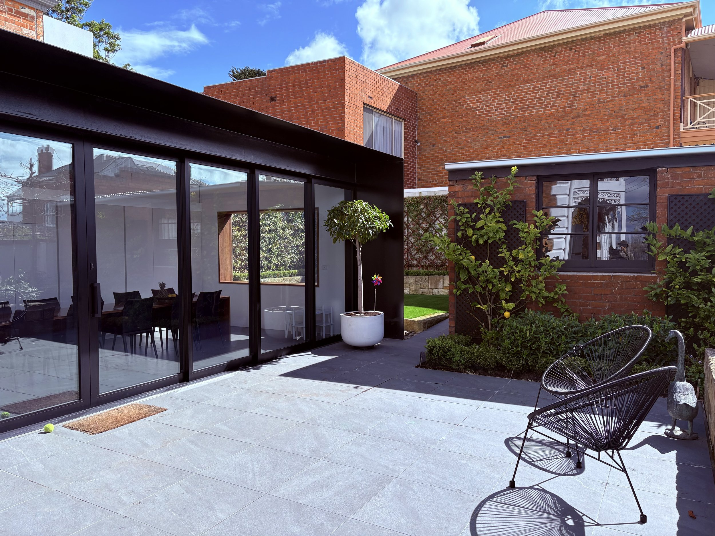 An outdoor patio area with large dark gray tiles, black wire chairs, a small round table, a potted tree, and a wind spinner, with a brick building and a glass-enclosed room reflecting the sky and other buildings in the background.