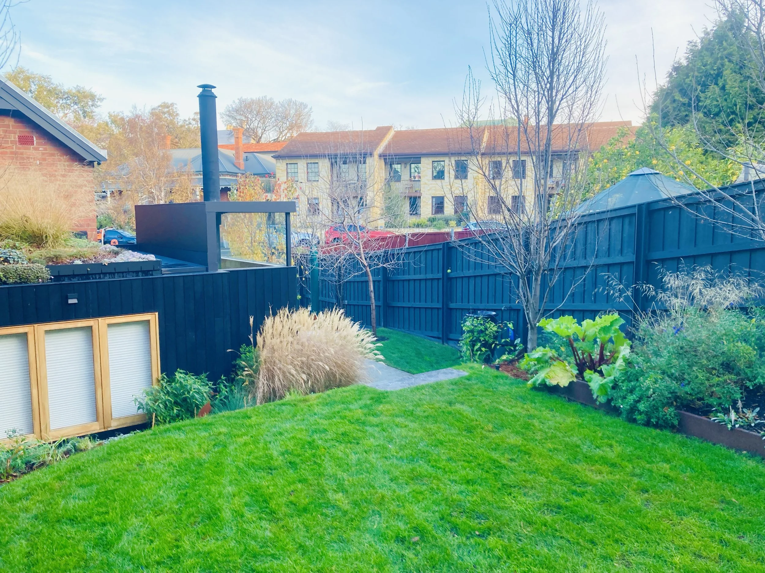 A backyard with a green lawn, trees, a black wooden fence, and a view of residential buildings. There is a chimney on the roof and a black outdoor fireplace or grill in the yard. The sky is clear and blue.