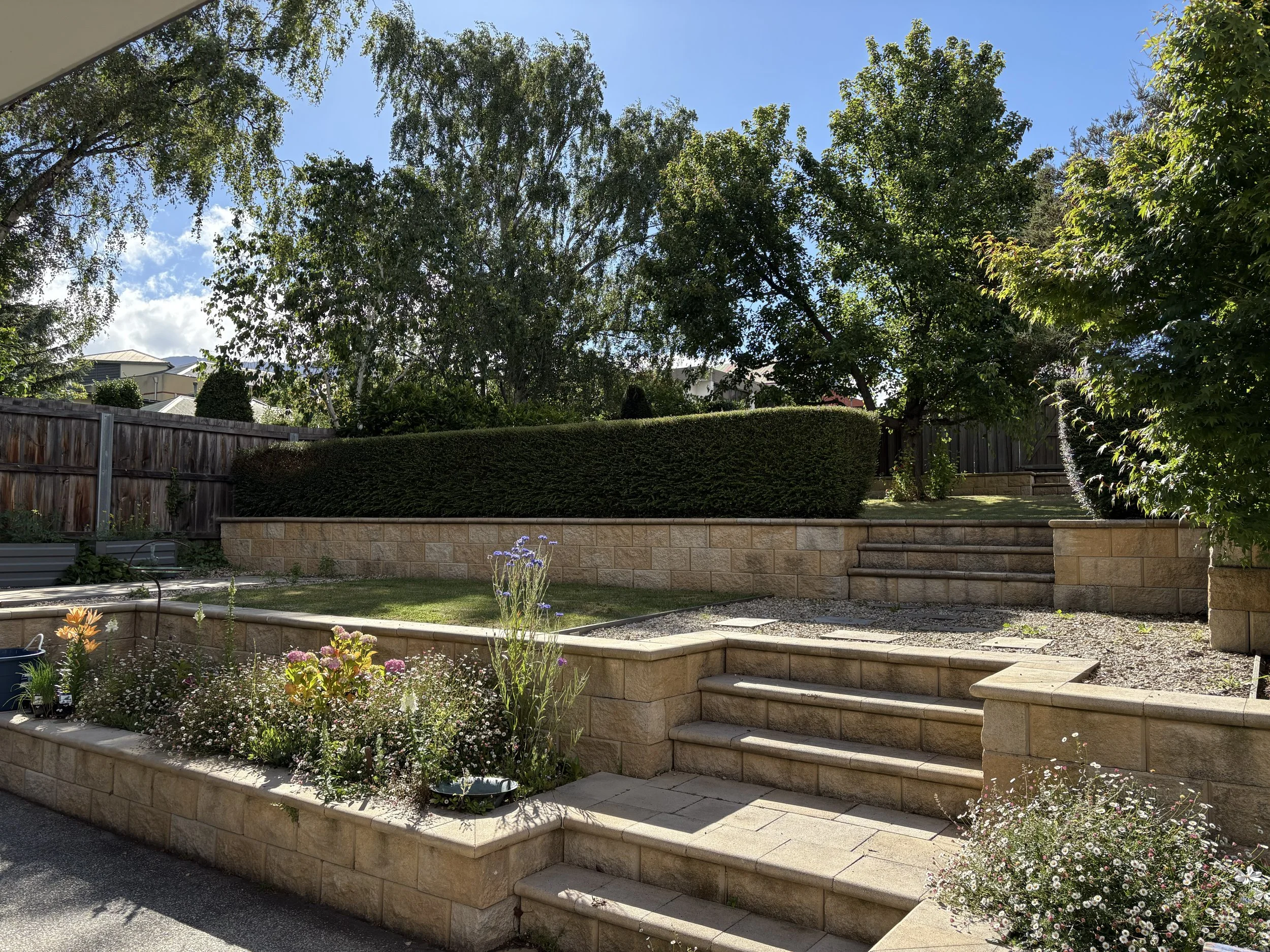 A backyard garden with stone steps leading up to a grassy area. The garden has flowerbeds with various colorful flowers and shrubs, bordered by a low brick wall. Tall trees and a wooden fence surround the yard, and the sky is clear with some clouds.