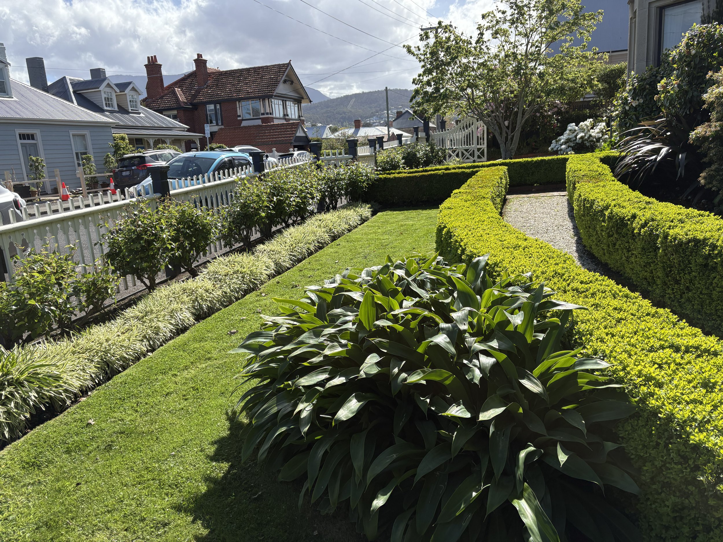A sunny residential neighborhood with a well-maintained garden featuring neatly trimmed hedges, a gravel pathway, and lush green plants. There are houses with pitched roofs, some brick and some wooden, parked cars along the street, and hills with tre