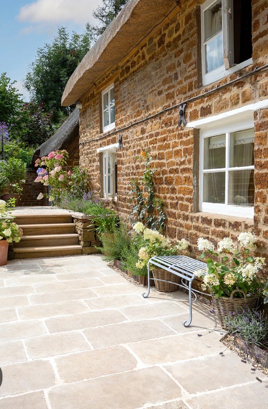 A brick house with white-framed windows, a stone pathway, and a garden with various flowers and plants. There is a small metal bench on the patio.