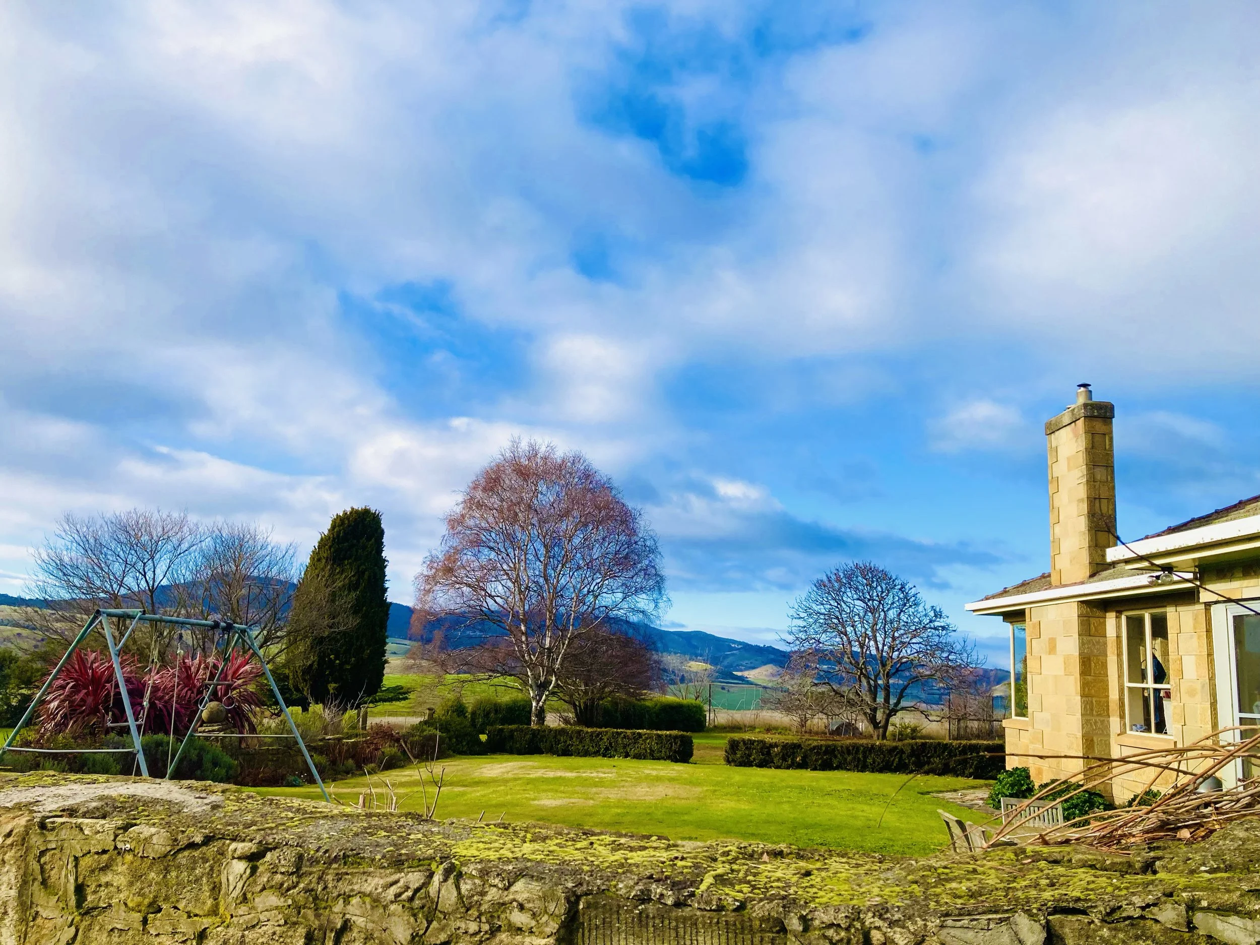 A backyard with a stone wall, green grass, and trees with no leaves. There is a swing set on the left and a house with a chimney on the right. In the background, there are hills and a cloudy sky.