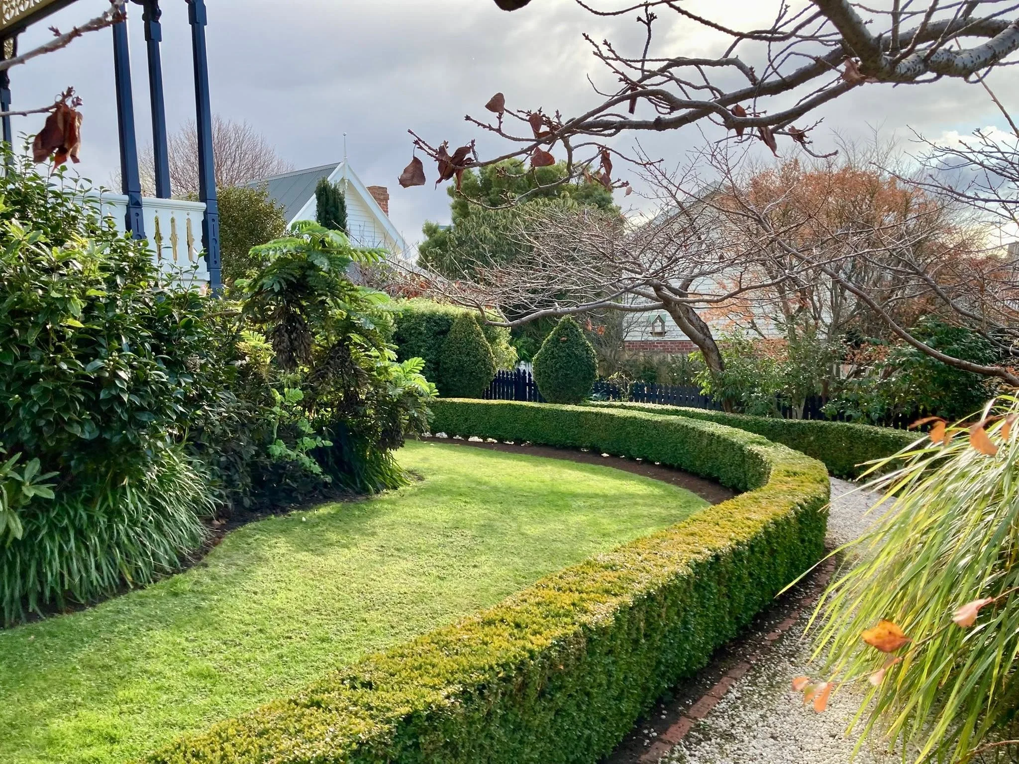 A well-manicured backyard garden with lush green grass, edged with hedges and various trees, with a house in the background under a cloudy sky.