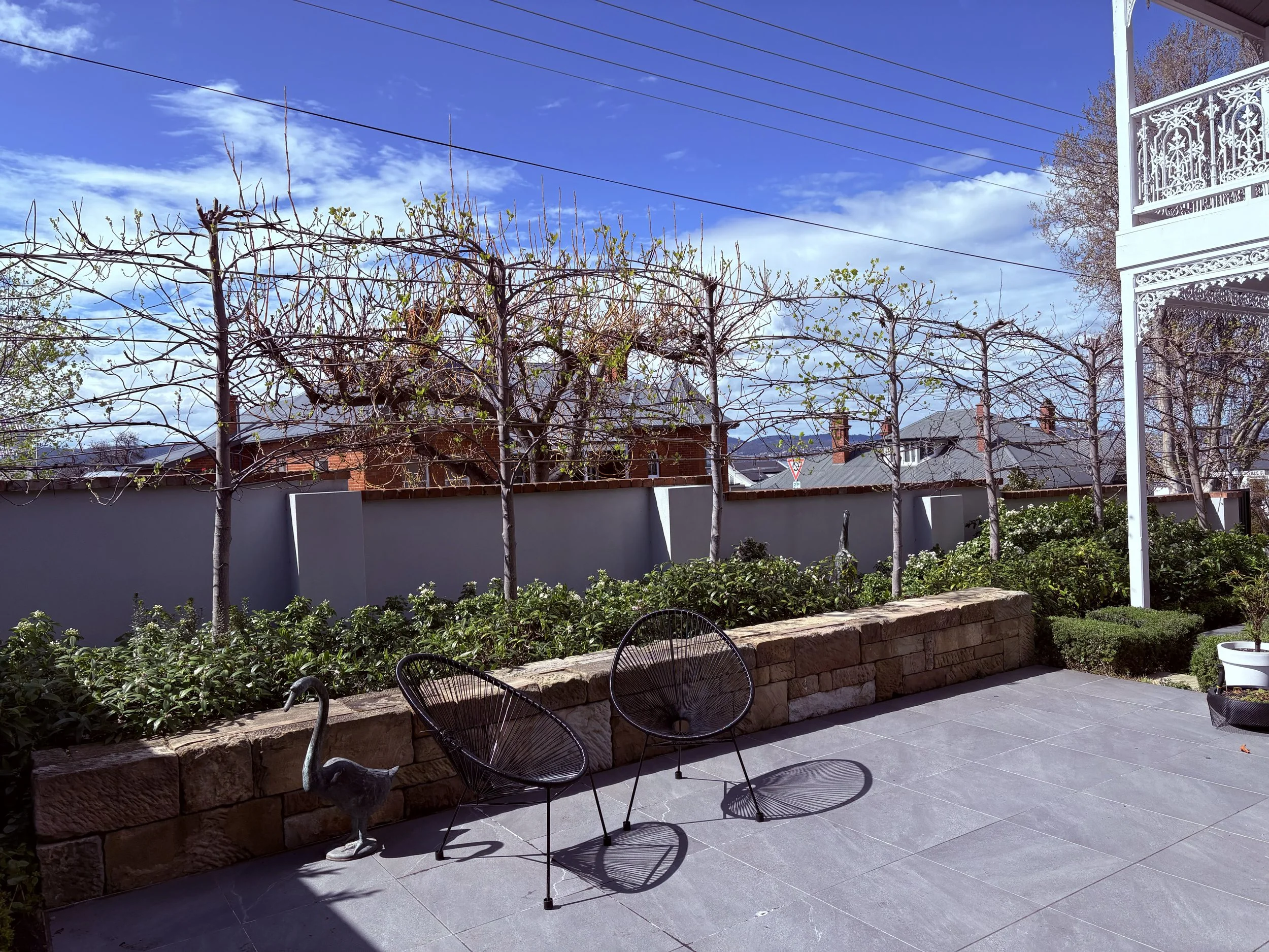 A backyard patio with a grey stone floor, a low stone wall, and a row of leafless trees with some small green leaves. There are black metal chairs, a round white pot with a small plant, and a tall white decorative railing on the right. The background