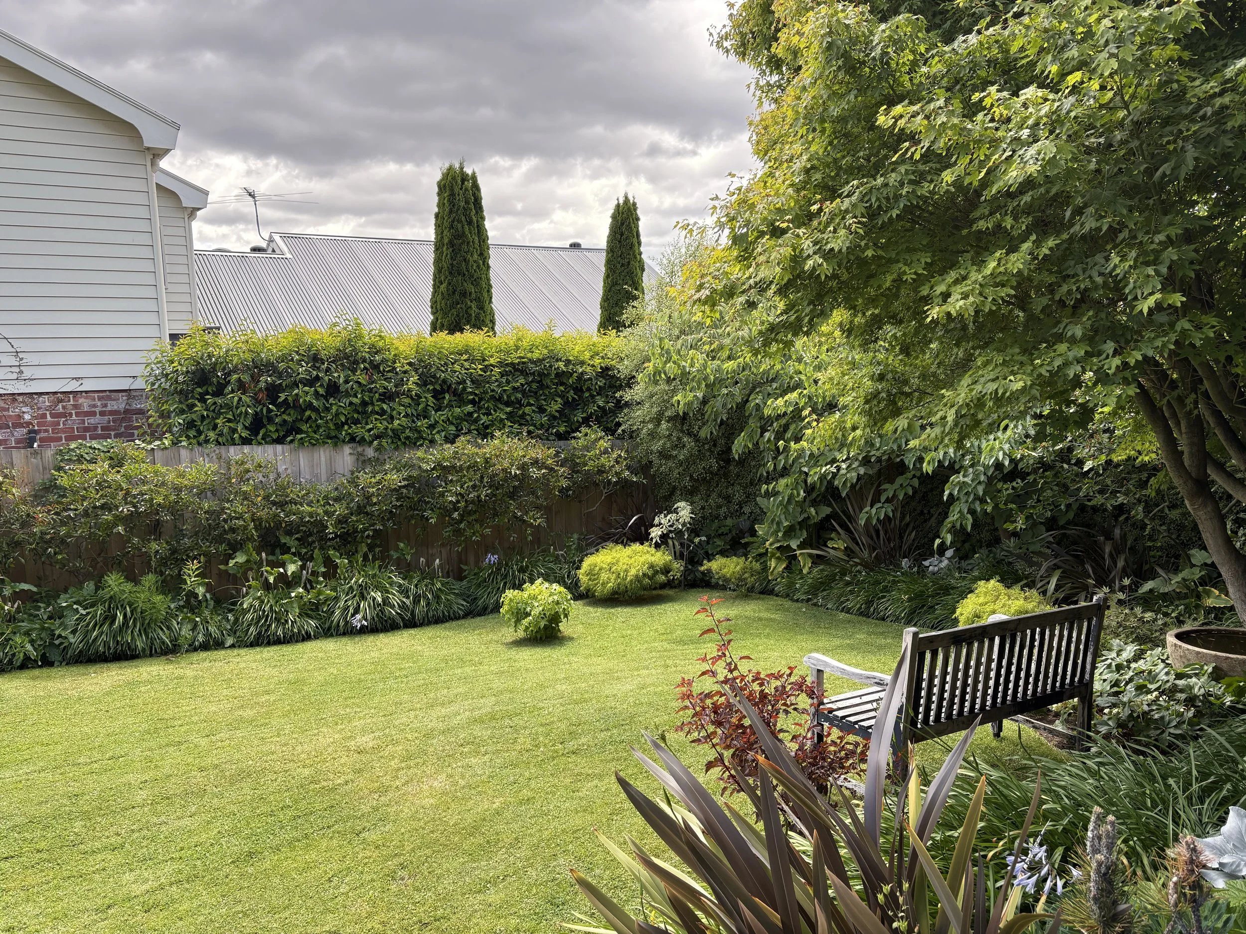 A cozy backyard garden with a well-maintained green lawn, various shrubs, and a bench. Tall trees and a hedge with lush green leaves surround the area, and a house with white siding and a metal roof is visible in the background. The sky is cloudy wit