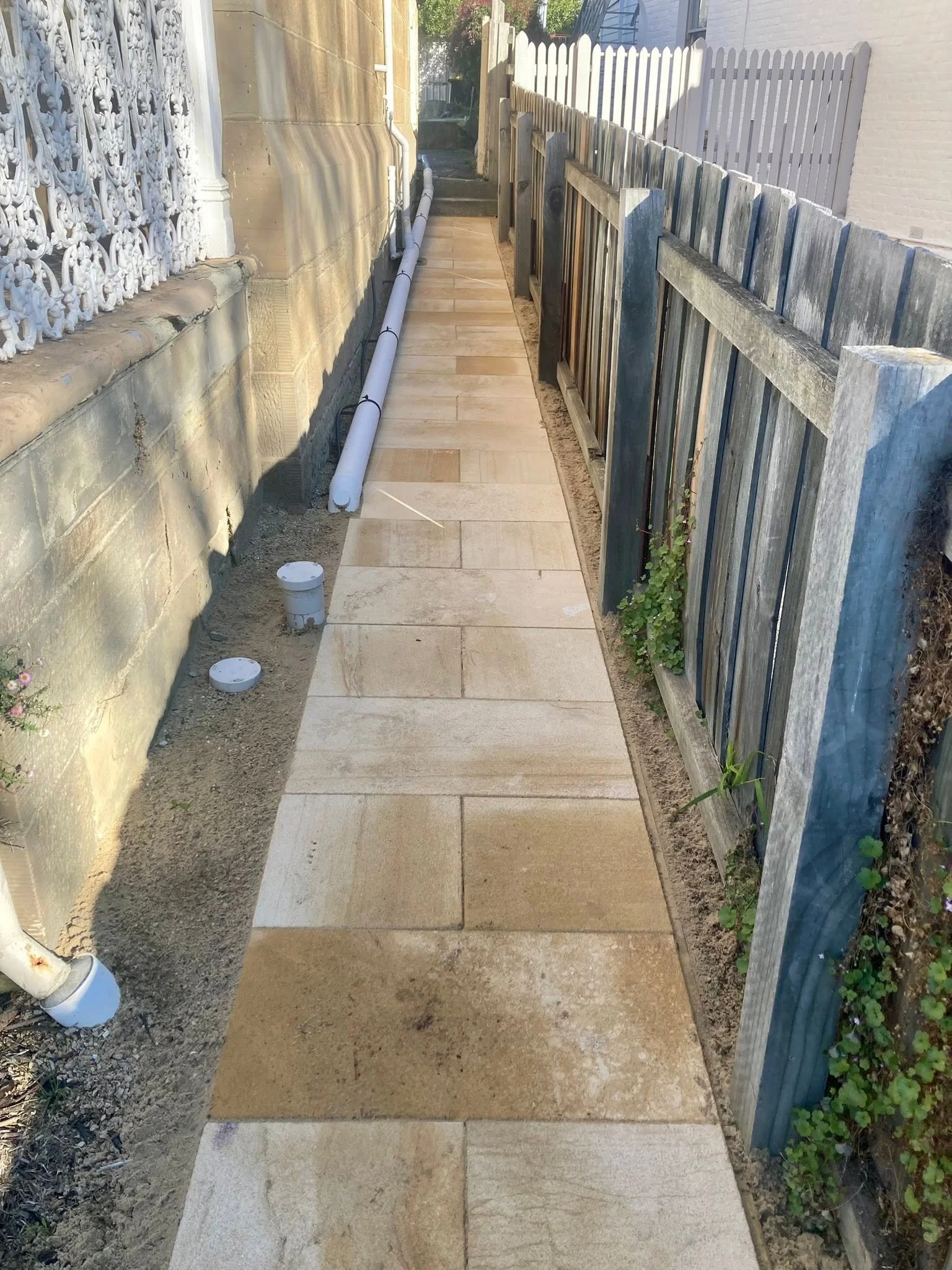 A narrow outdoor pathway laid with light-colored stone tiles, bordered on the left by a stone wall and on the right by a wooden fence, with construction pipes and small plants along the edges.