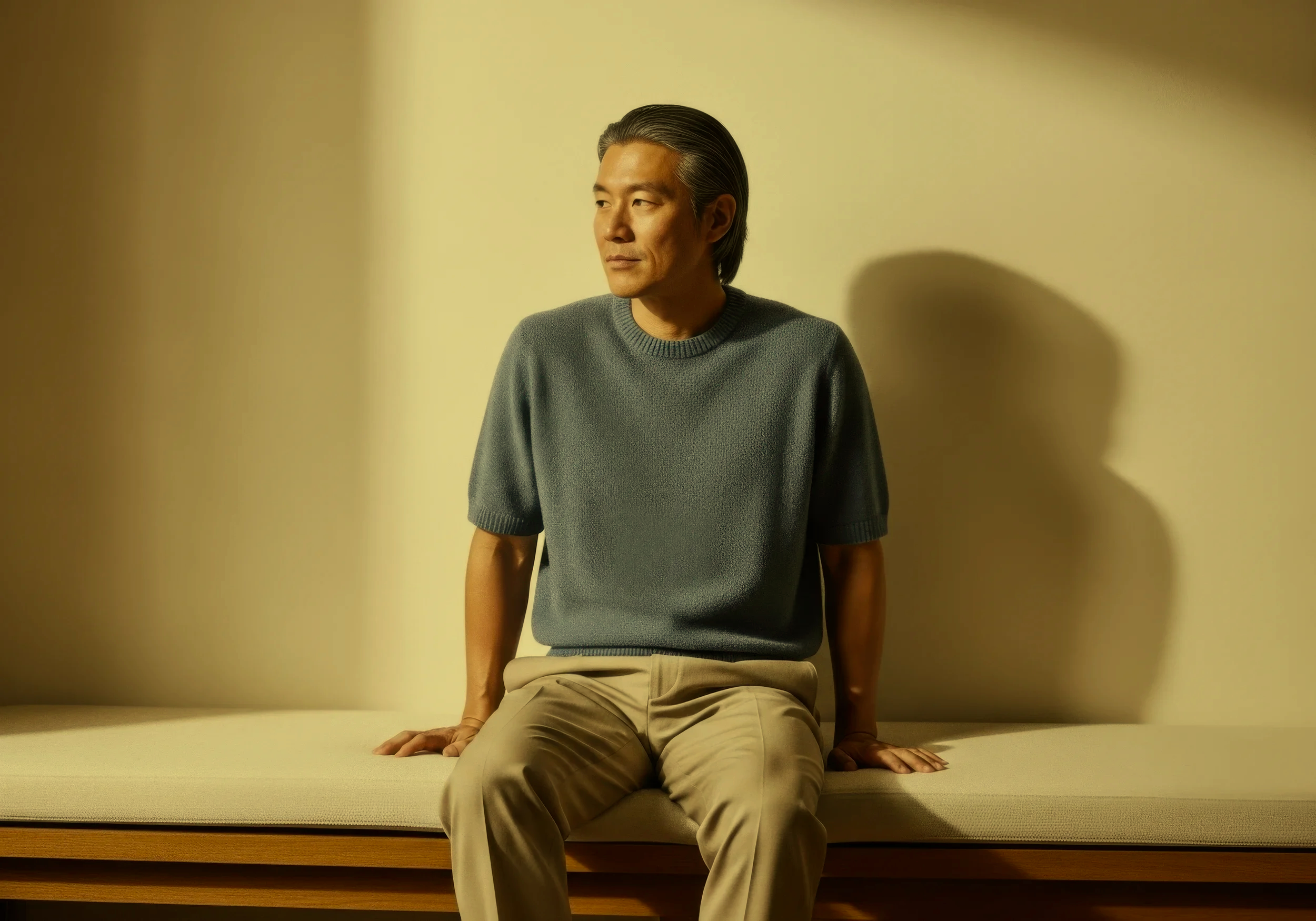 A middle-aged man with gray hair, wearing a gray sweater and beige pants, is sitting on a bench in front of a plain yellow wall. He is looking to his left with a calm expression, his left hand resting on the bench and his right hand also on the bench. His shadow is cast on the wall behind him.