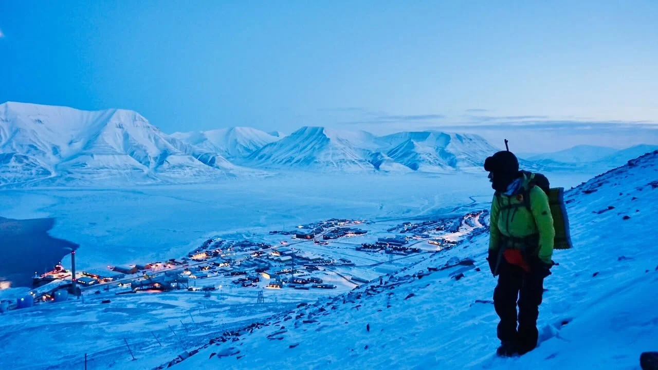 A person dressed in winter gear hiking on a snowy hillside overlooking a small town in a frozen landscape, with snow-covered mountains in the background at dusk or dawn.