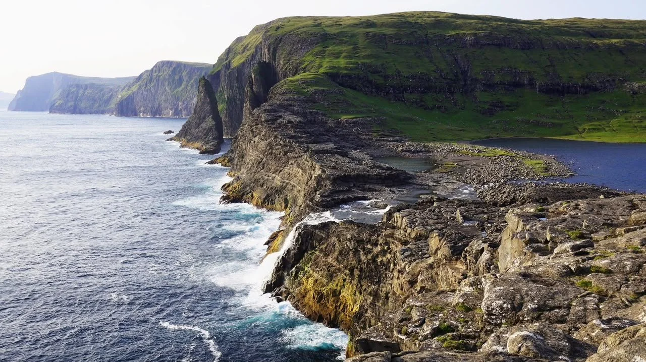 Cliffside landscape with green grassy slopes, rocky shoreline, and ocean waves crashing against the rocks.