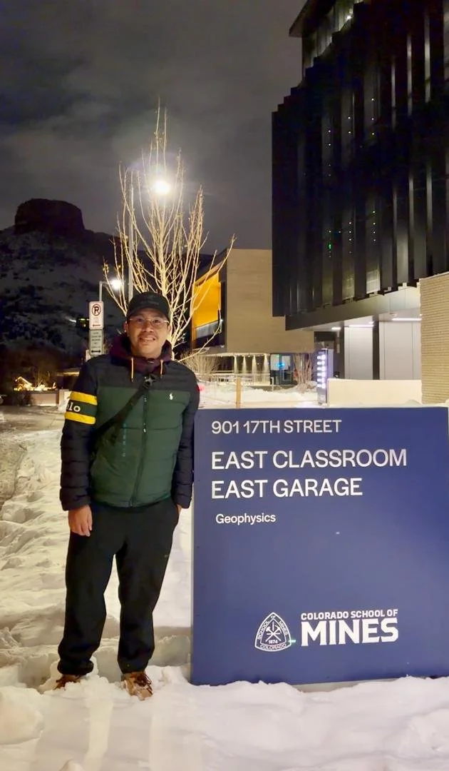 A man standing outdoors in snow at night, wearing a dark jacket, black cap, and glasses, next to a sign for the Colorado School of Mines. The sign indicates directions to the East Classroom and East Garage at 901 17th Street, and mentions geophysics.