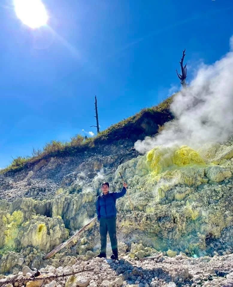 A person standing on rocky terrain holding a stick, with yellow sulfur deposits and fumaroles emitting steam in a volcanic area under a bright sun and blue sky.