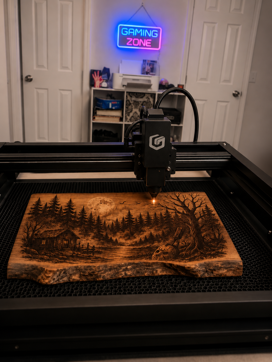 A laser engraving machine creating a detailed landscape scene on a wooden plaque featuring trees, a cabin, a moon, and a mask.