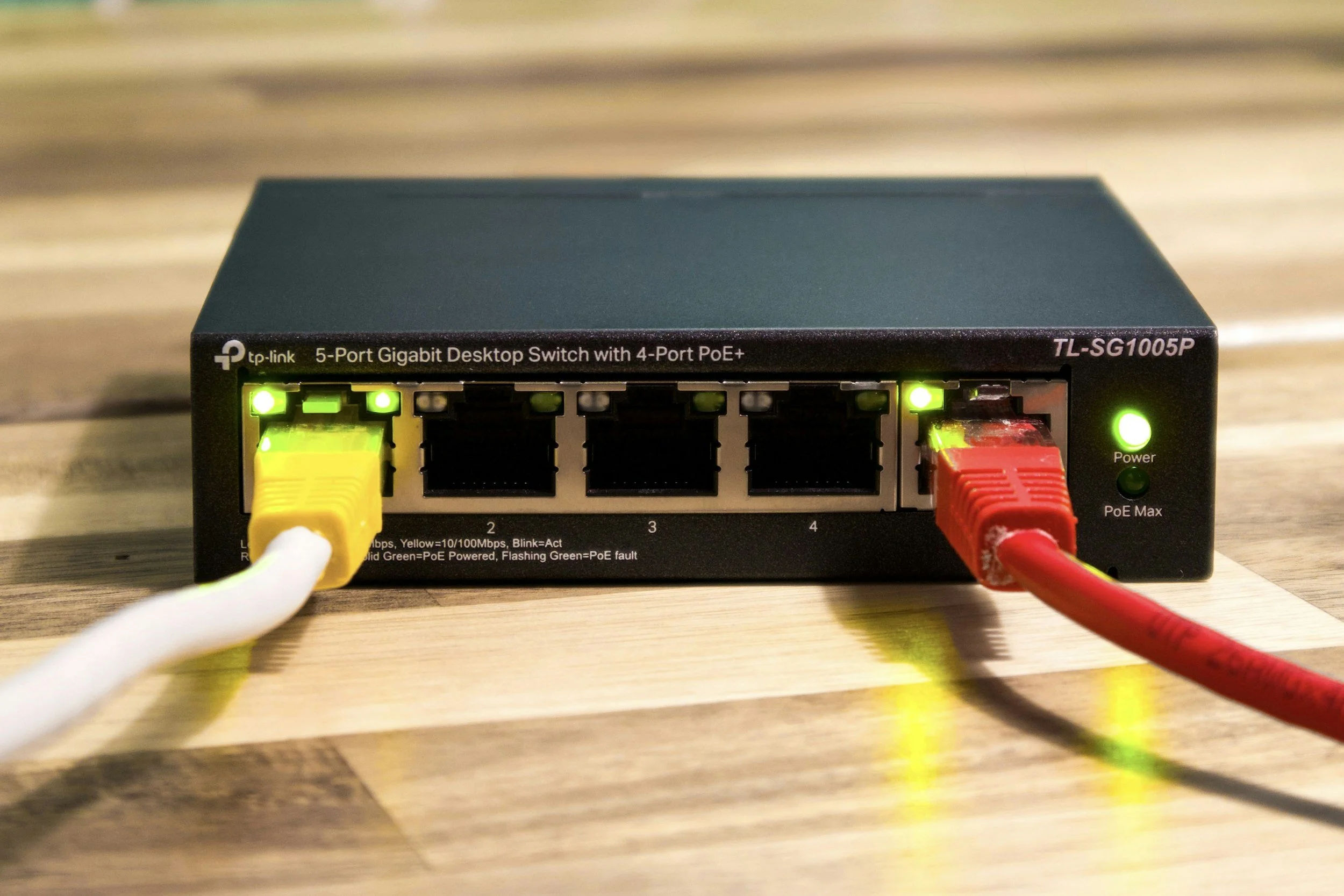 A network switch with multiple Ethernet cables connected to it, placed on a wooden surface. The switch has indicator lights and is labeled 'tp-link 5-Port Gigabit Desktop Switch with 4-Port PoE+.'