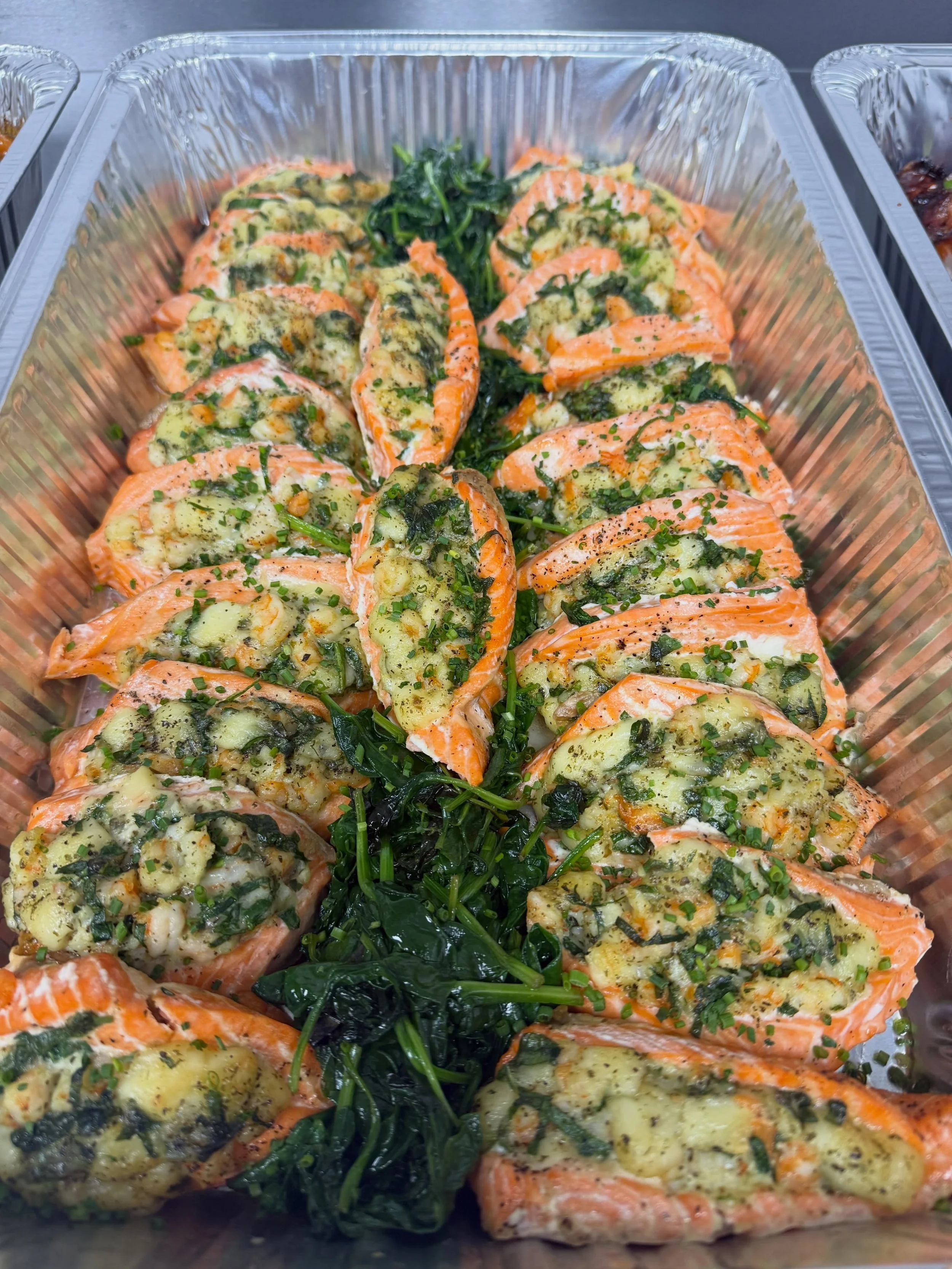 Baked salmon fillets topped with herbs and garlic, served with sautéed greens in a foil tray.