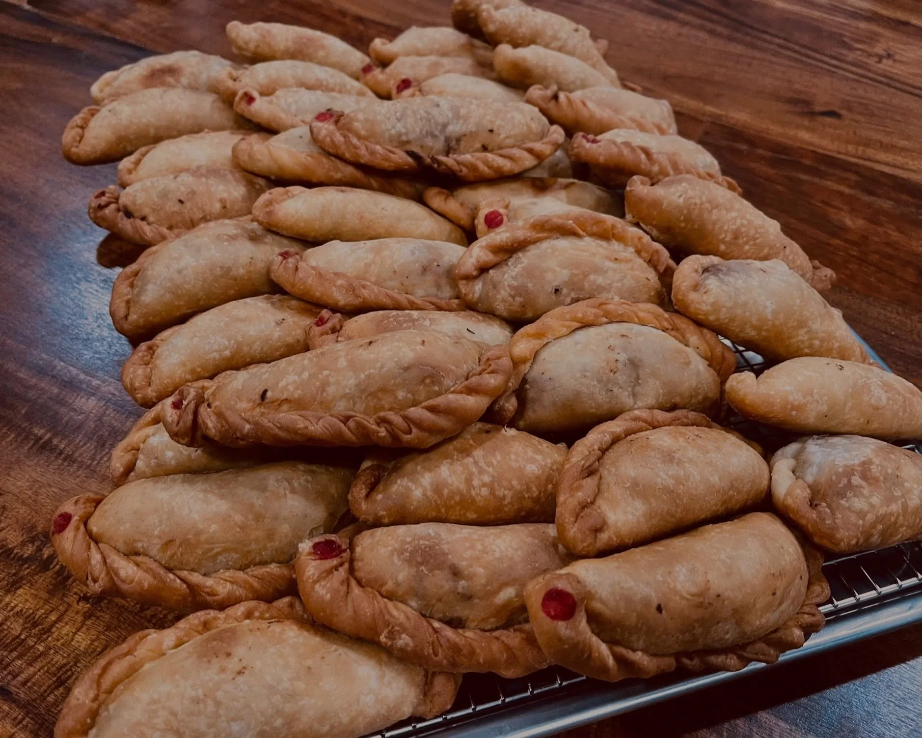 A tray of freshly baked empanadas with crimped edges and a golden-brown crust, some with red spots, sitting on a wire rack on a wooden surface.