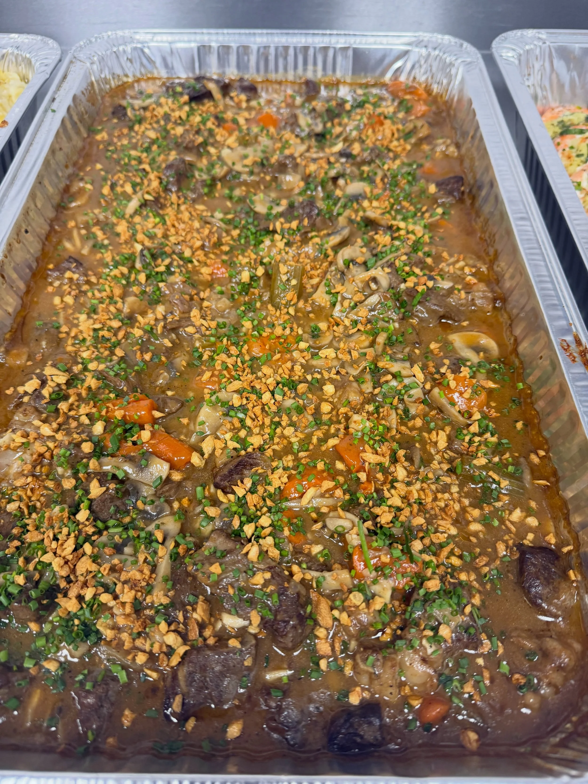 A large aluminum tray filled with beef stew containing chunks of beef, vegetables, and topped with chopped green onions and crushed crackers.