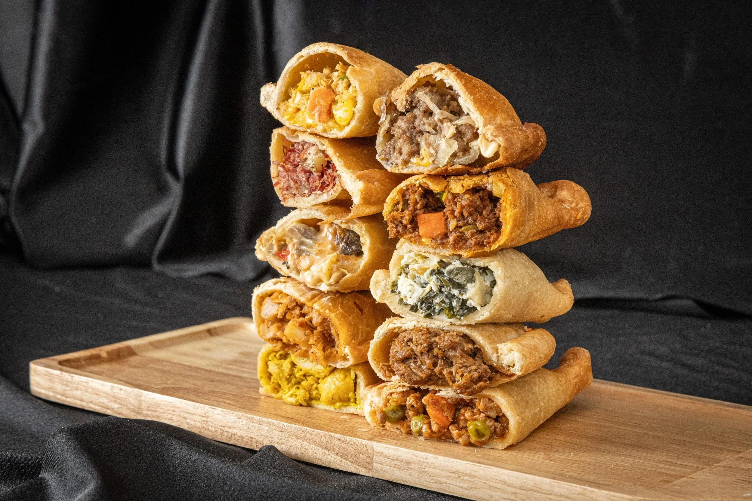 Stacked egg rolls filled with various savory ingredients on a wooden serving board, with a dark background.