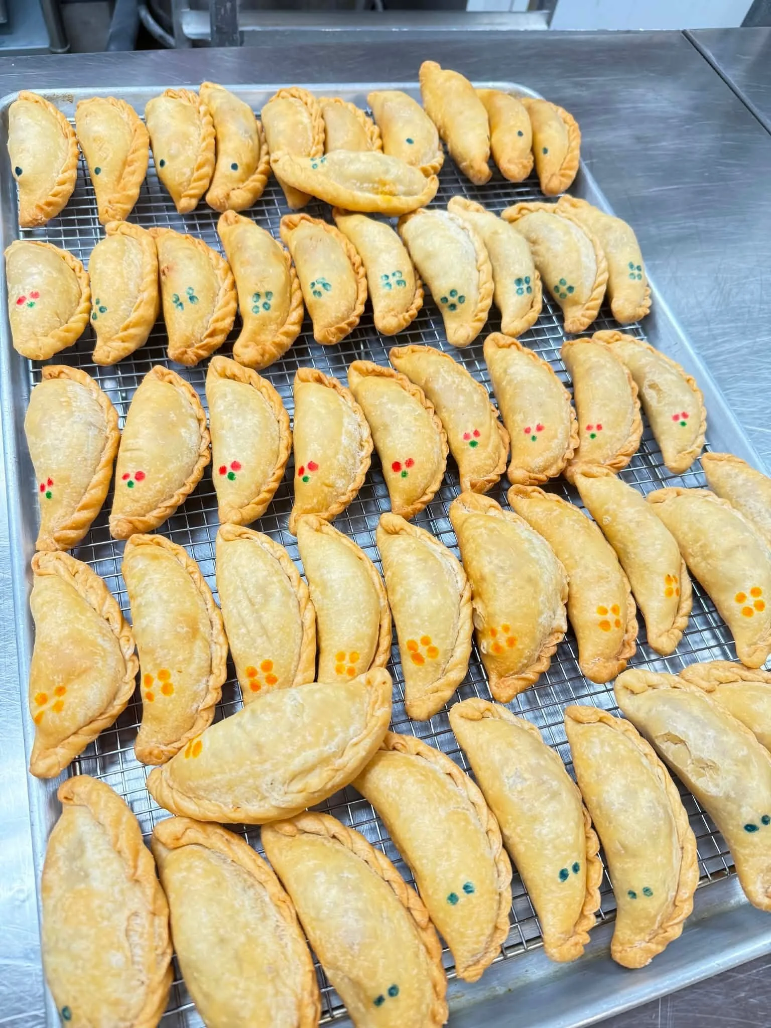 A tray of baked empanadas with small colored decorative dots on some of them, placed on a metal countertop.