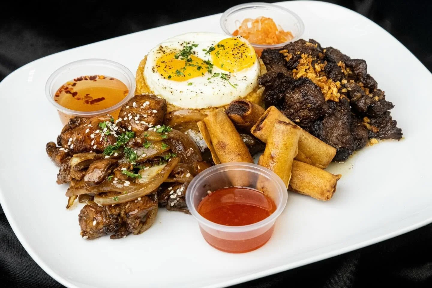 Plate with sunny side up eggs, tamales, fried beef, caramelized onions with sesame seeds, and small containers of salsa and orange sauce