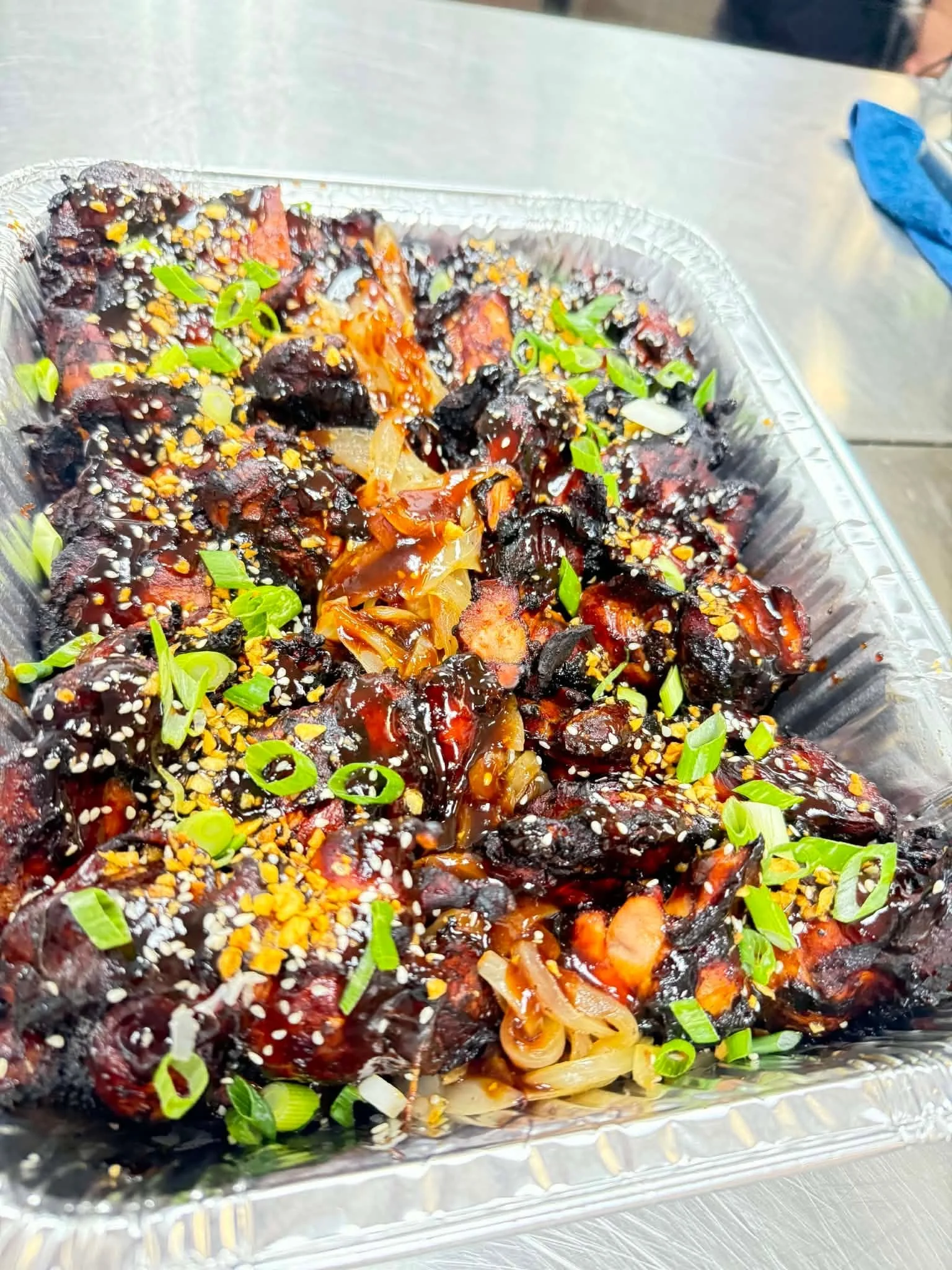 Barbecue pork ribs garnished with chopped green onions and sesame seeds in a foil tray.