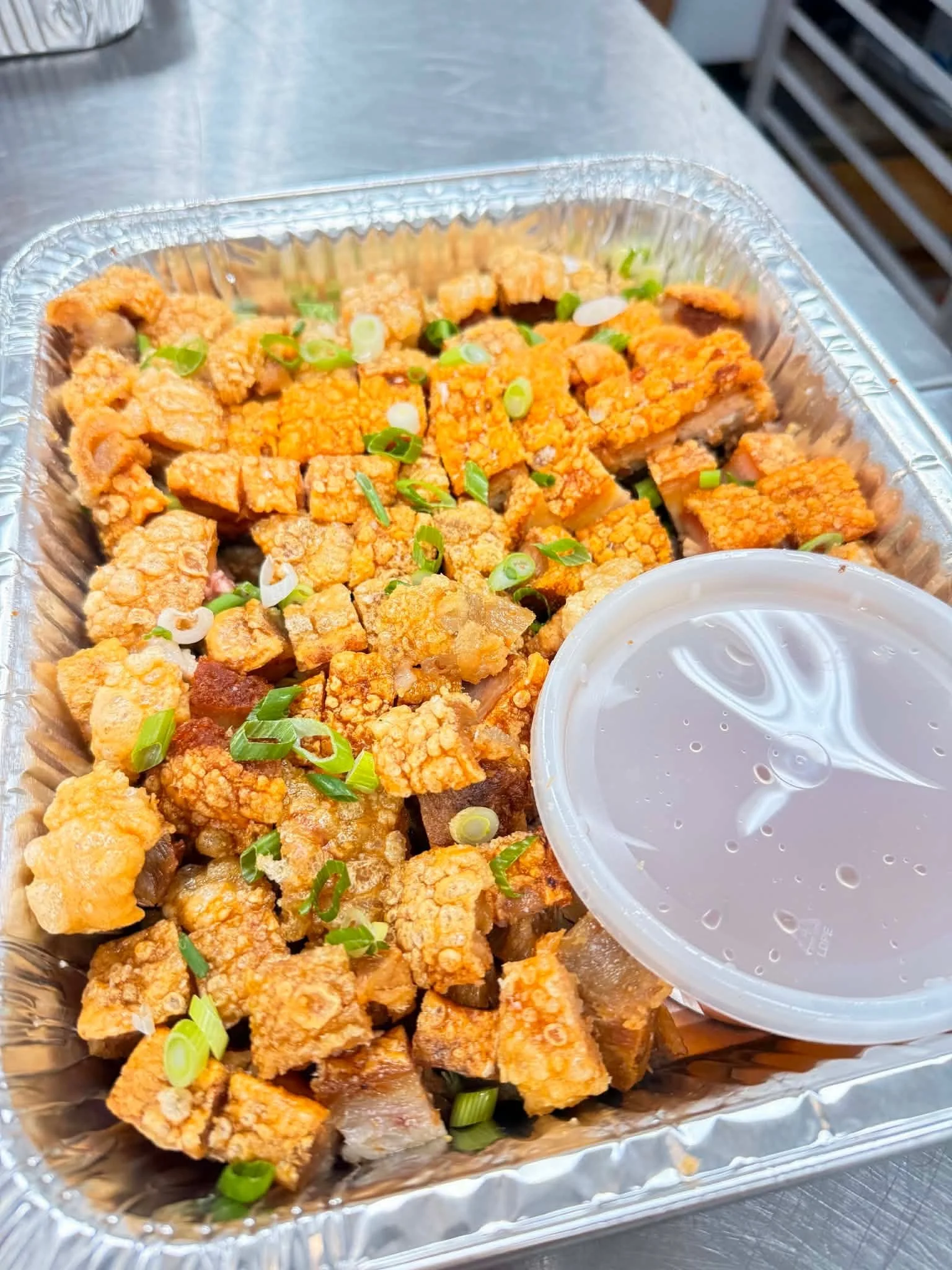 Fried pieces of chicken garnished with chopped green onions in a foil container, with a cup of dipping sauce on the side.