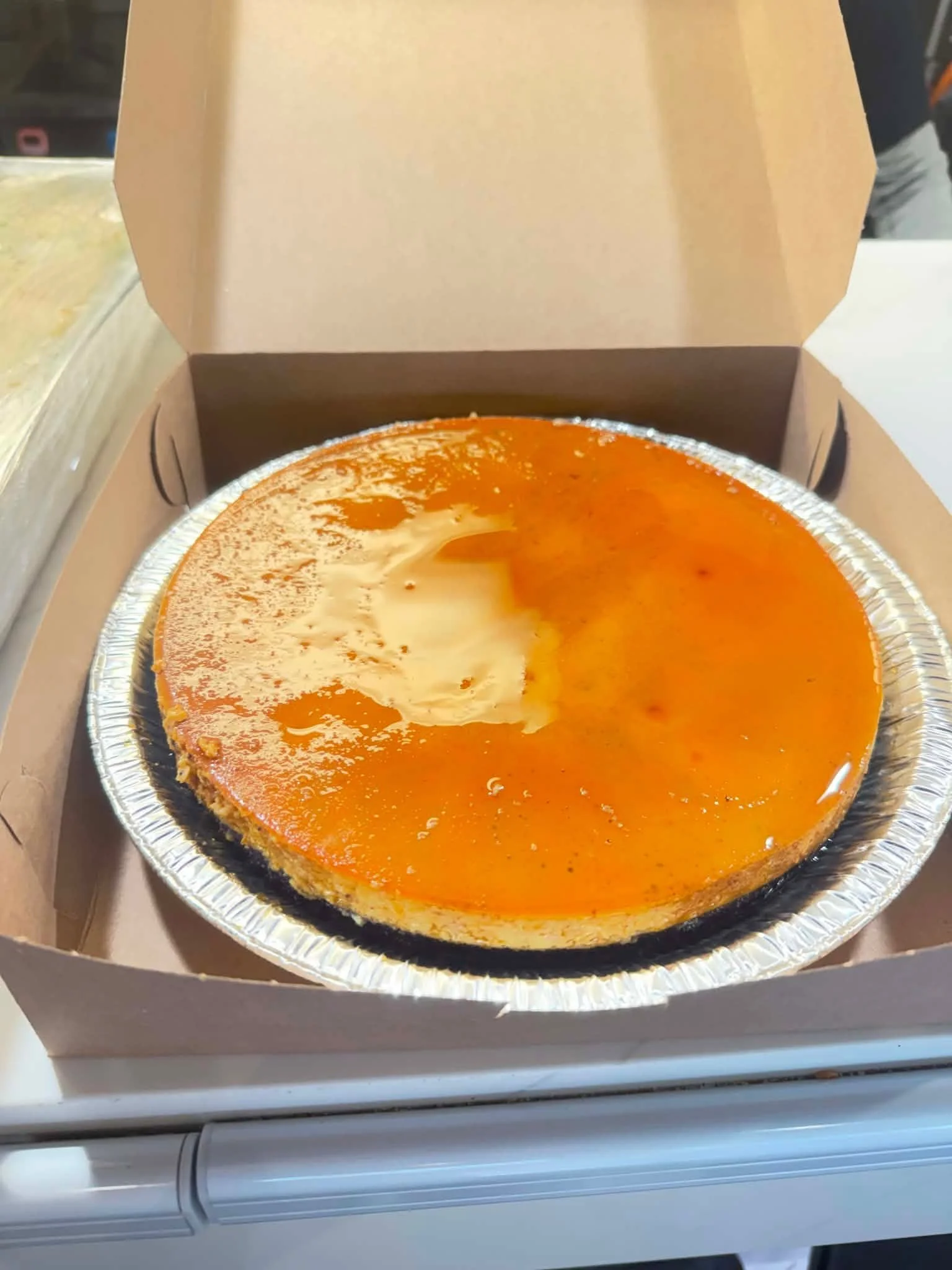 A pumpkin cheesecake in a foil pan inside a cardboard box, sitting on a white surface.