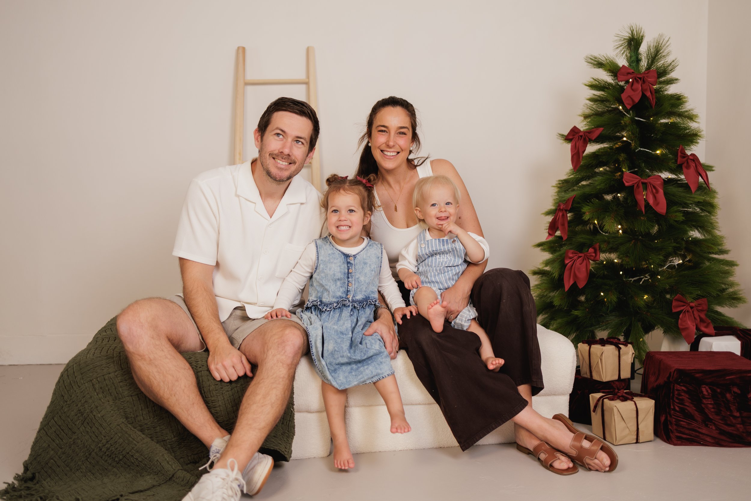 A family of four sitting on a white couch with a decorated Christmas tree beside them, surrounded by wrapped presents. The family is smiling, with two young daughters and their parents, in a cozy indoor setting.
