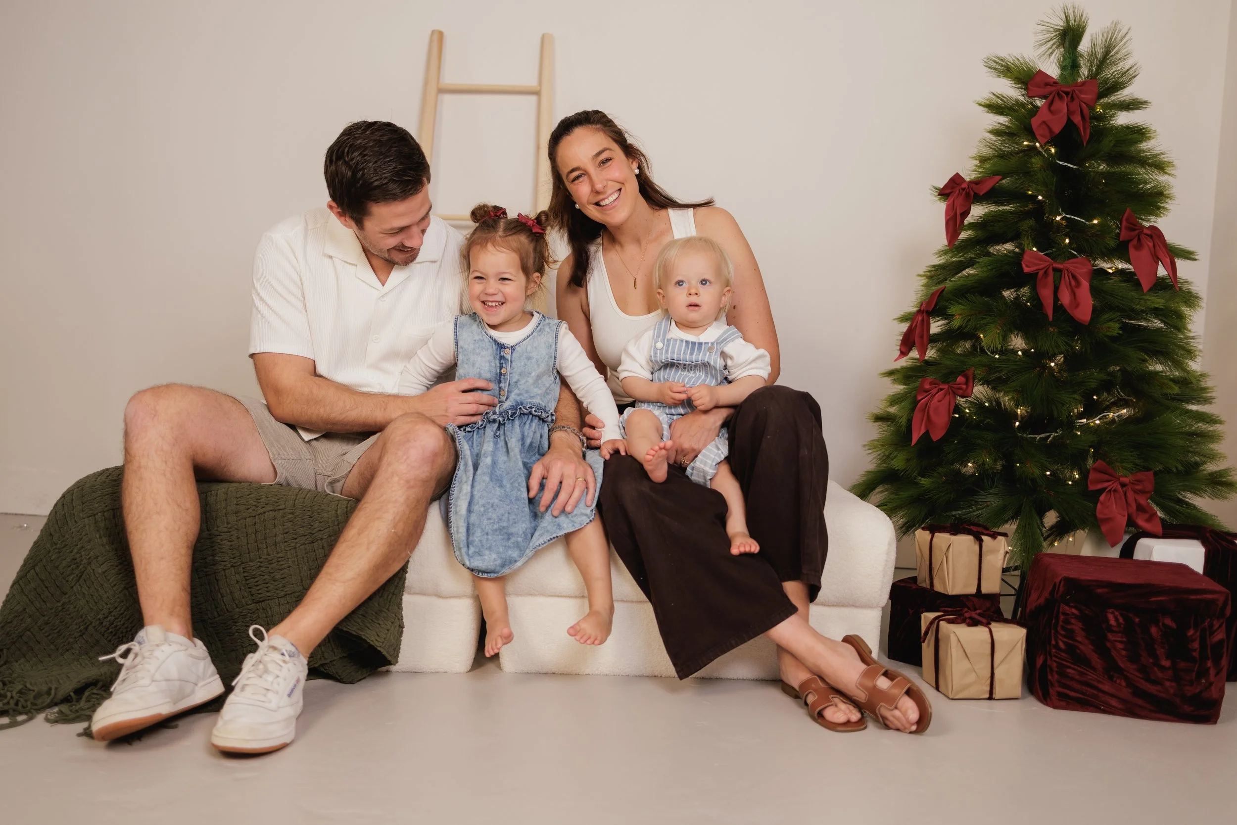 A family of four sitting on a white sofa near a decorated Christmas tree with gifts underneath, smiling and enjoying the moment.