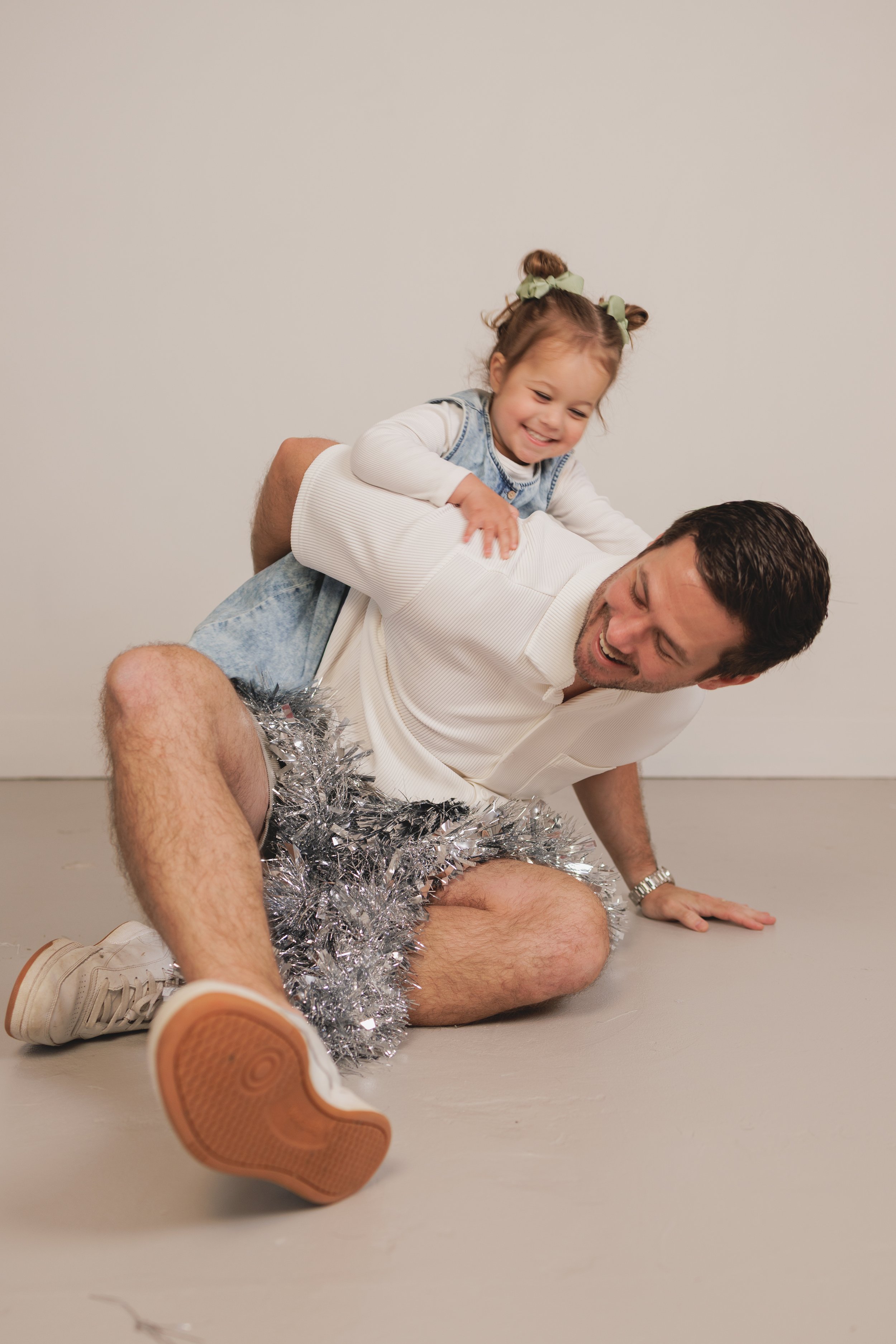A man on the floor with a young girl on his back, both smiling. The girl has two green hair ribbons and pigtails, and the man is wearing a white shirt. There is silver tinsel around the man's legs, and the background is plain and light-colored.