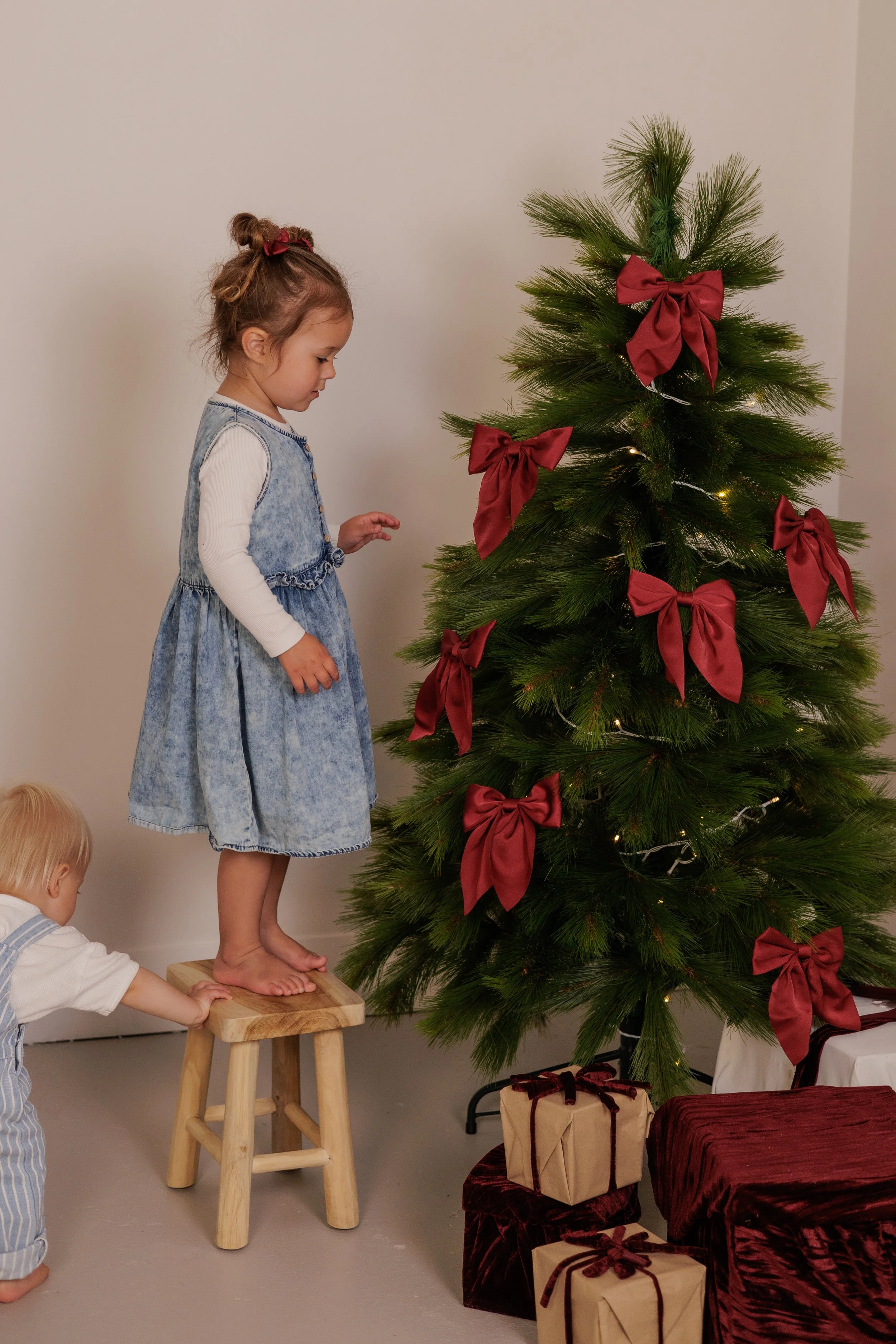 A young girl in a denim dress standing on a wooden stool, looking at a decorated Christmas tree with red bows and wrapped presents underneath. Another small child is reaching for the stool.