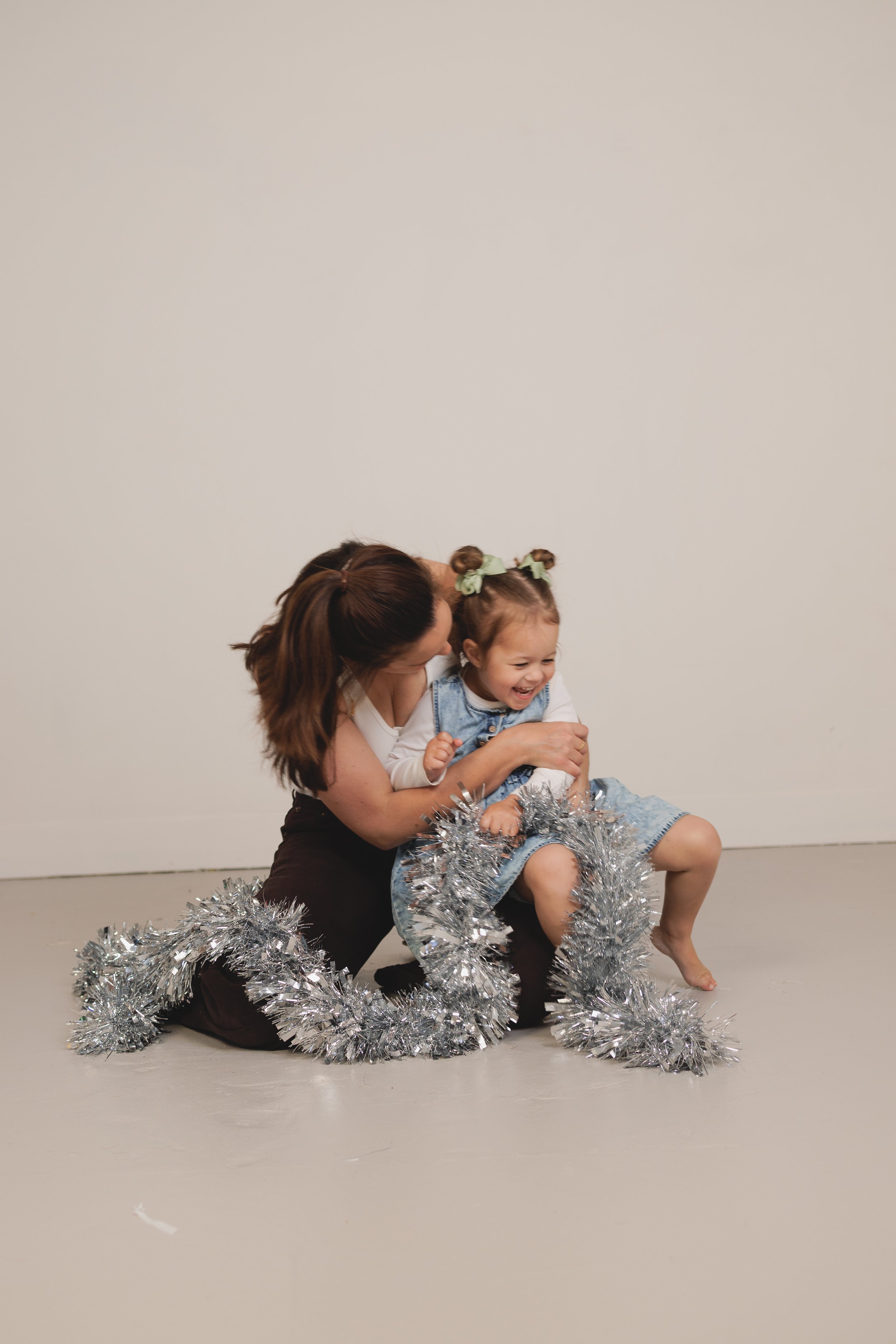 A woman and a young girl sitting on the ground surrounded by silver tinsel, with the woman hugging the girl and both smiling.