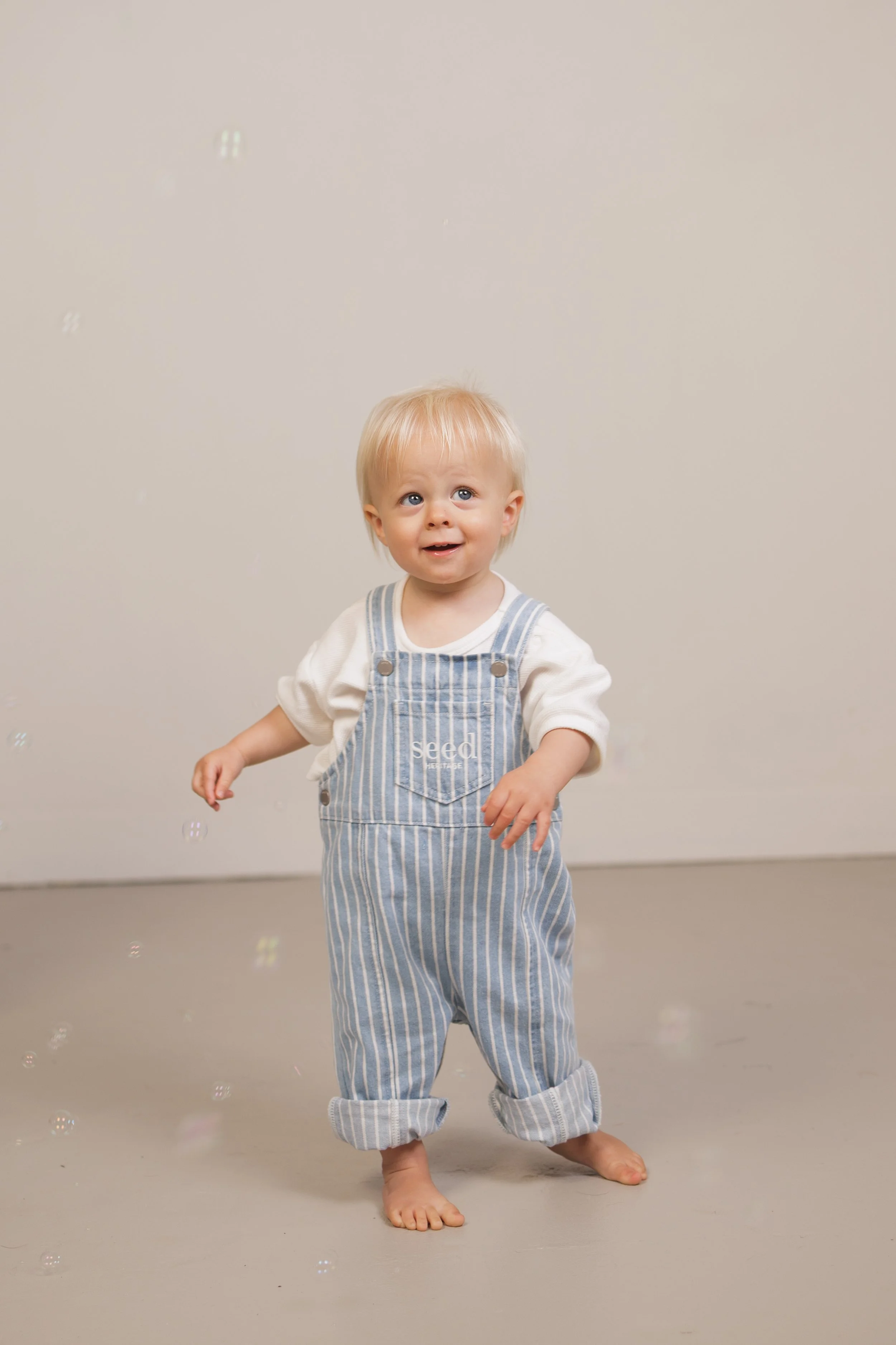 A young child with blond hair and blue eyes is standing barefoot on a light gray floor against a plain beige wall, looking up with a happy expression. The child is wearing a white shirt under striped overalls with the name 'Seed' on the front pocket.