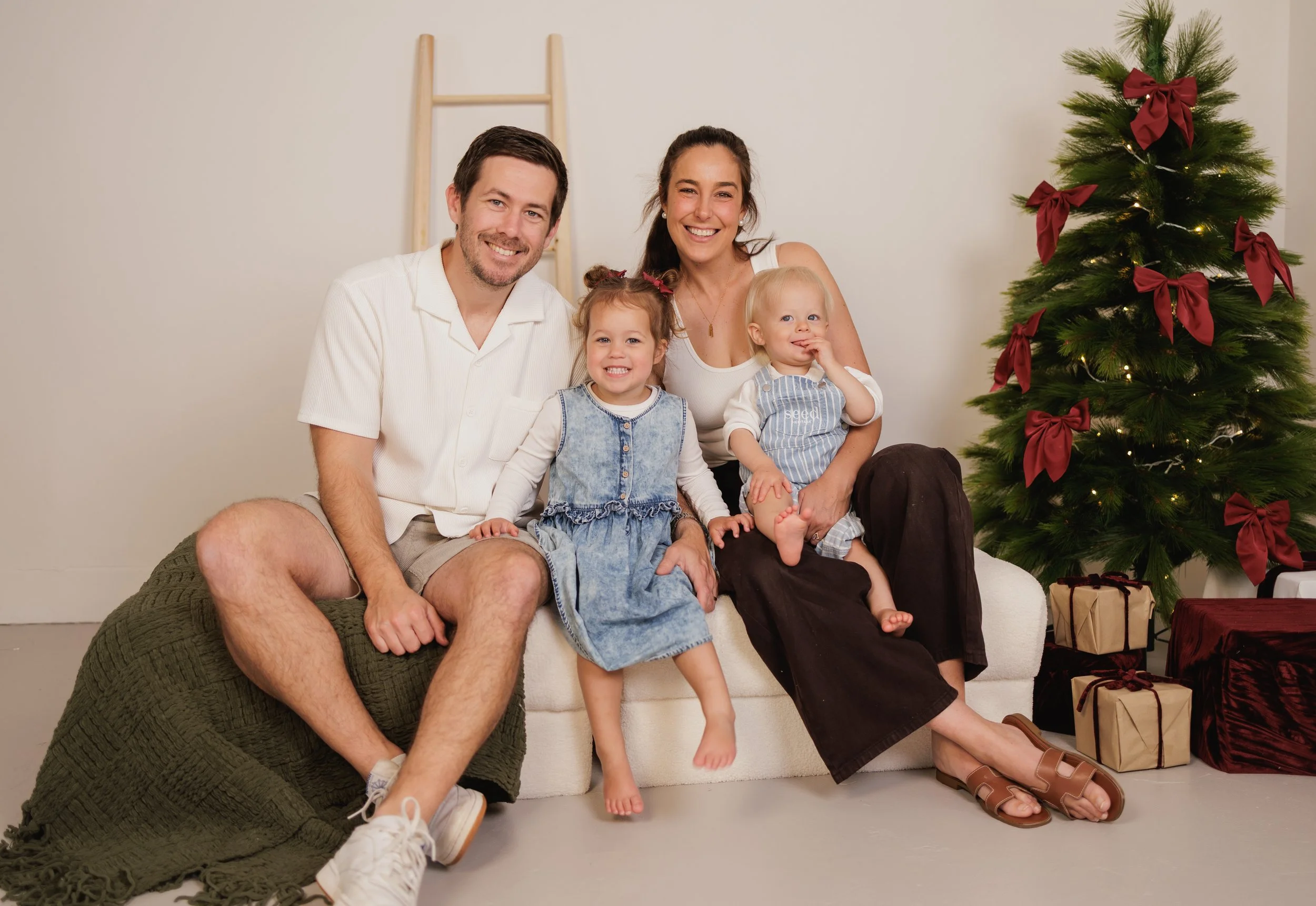 A family of four, including a father, mother, and their two young daughters, sits on a white sofa in front of a decorated Christmas tree with red bows and presents, smiling indoors.