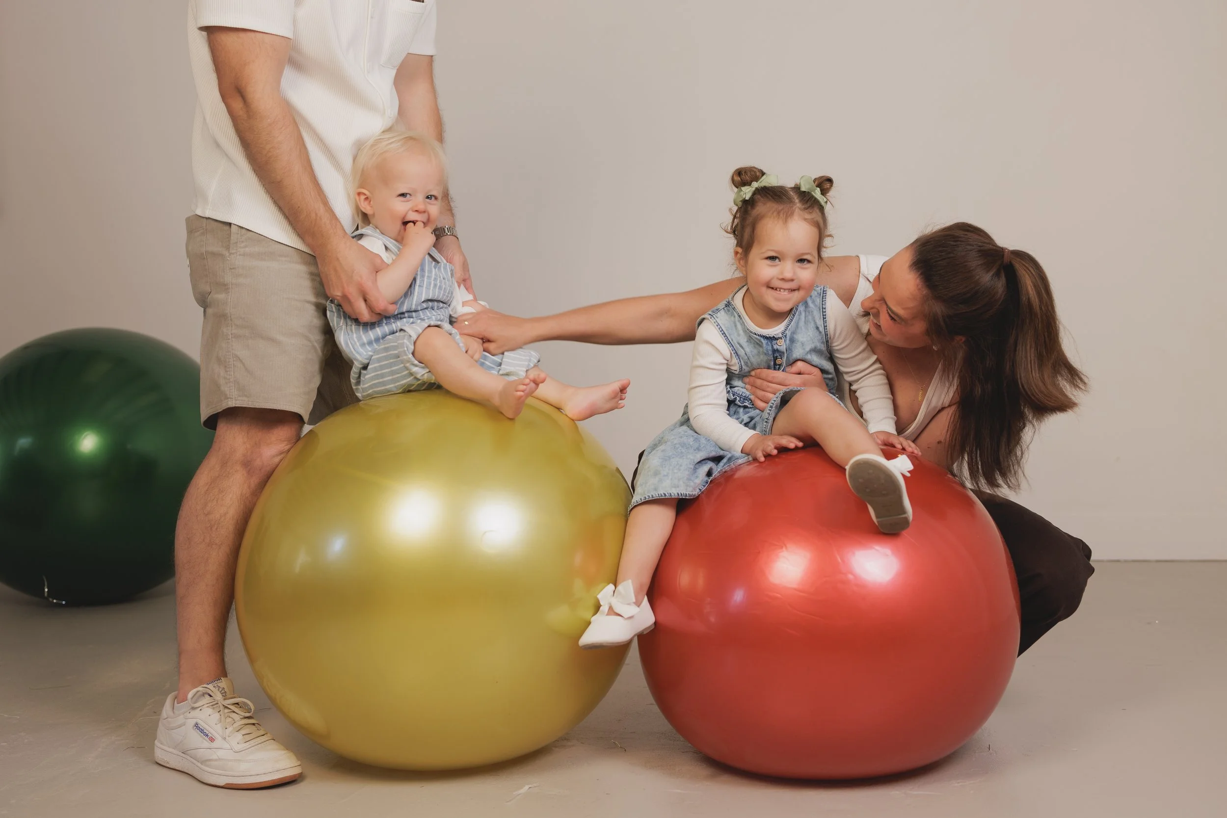 Two young girls sitting on large exercise balls, smiling, with a woman leaning over them and a man standing behind them. Two smaller exercise balls are on the floor nearby.