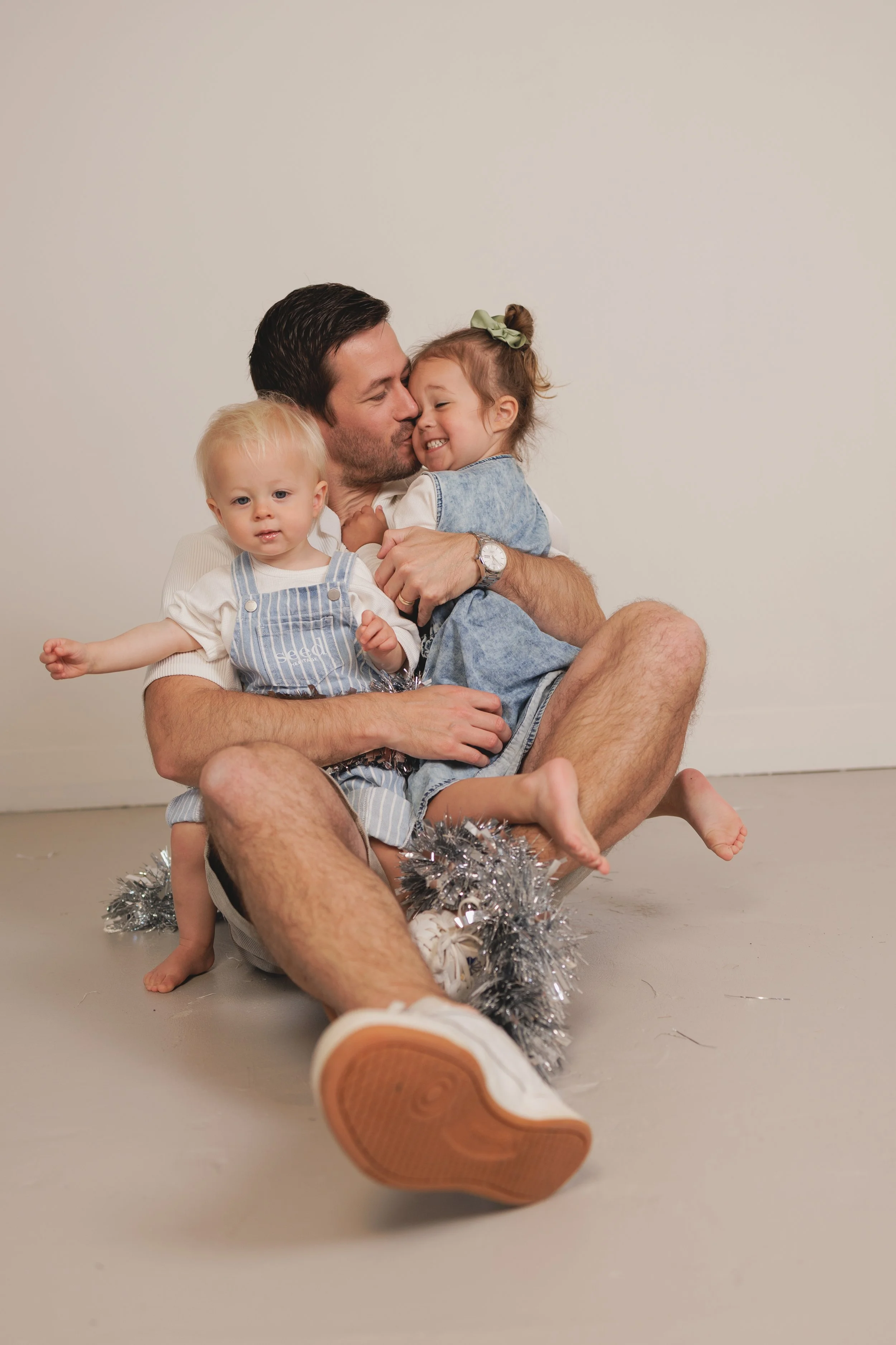 A man with two young children sitting on the floor with a white background. The man is holding a girl close to him, smiling and kissing her cheek. The boy sitting on his lap looks at the camera with a neutral expression. There is silver tinsel on the