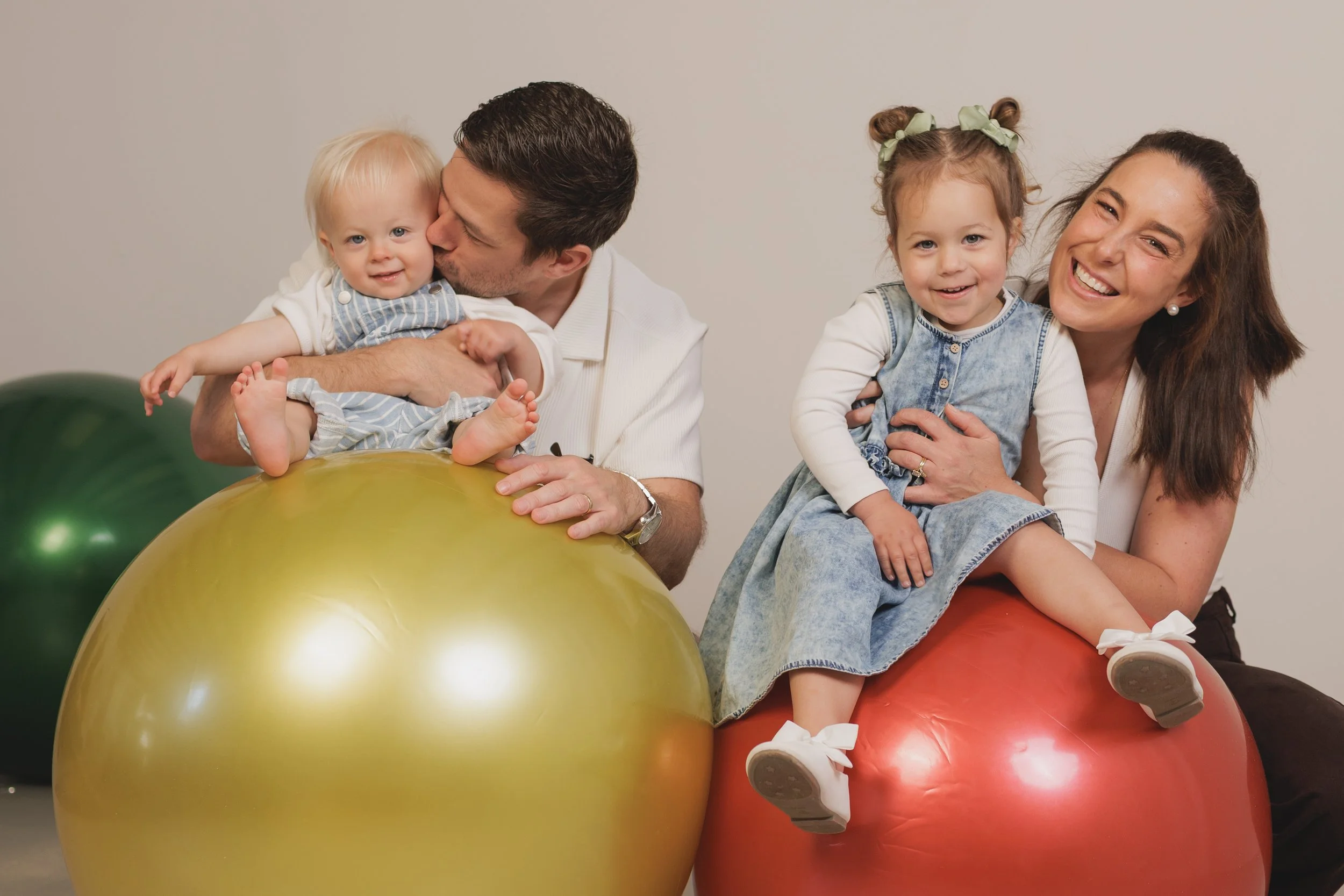A family of four sitting on large red, yellow, and green exercise balls, smiling and enjoying each other's company against a plain background.